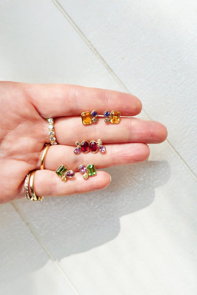 Hand holding multiple pairs of colorful gemstone earrings on a white background