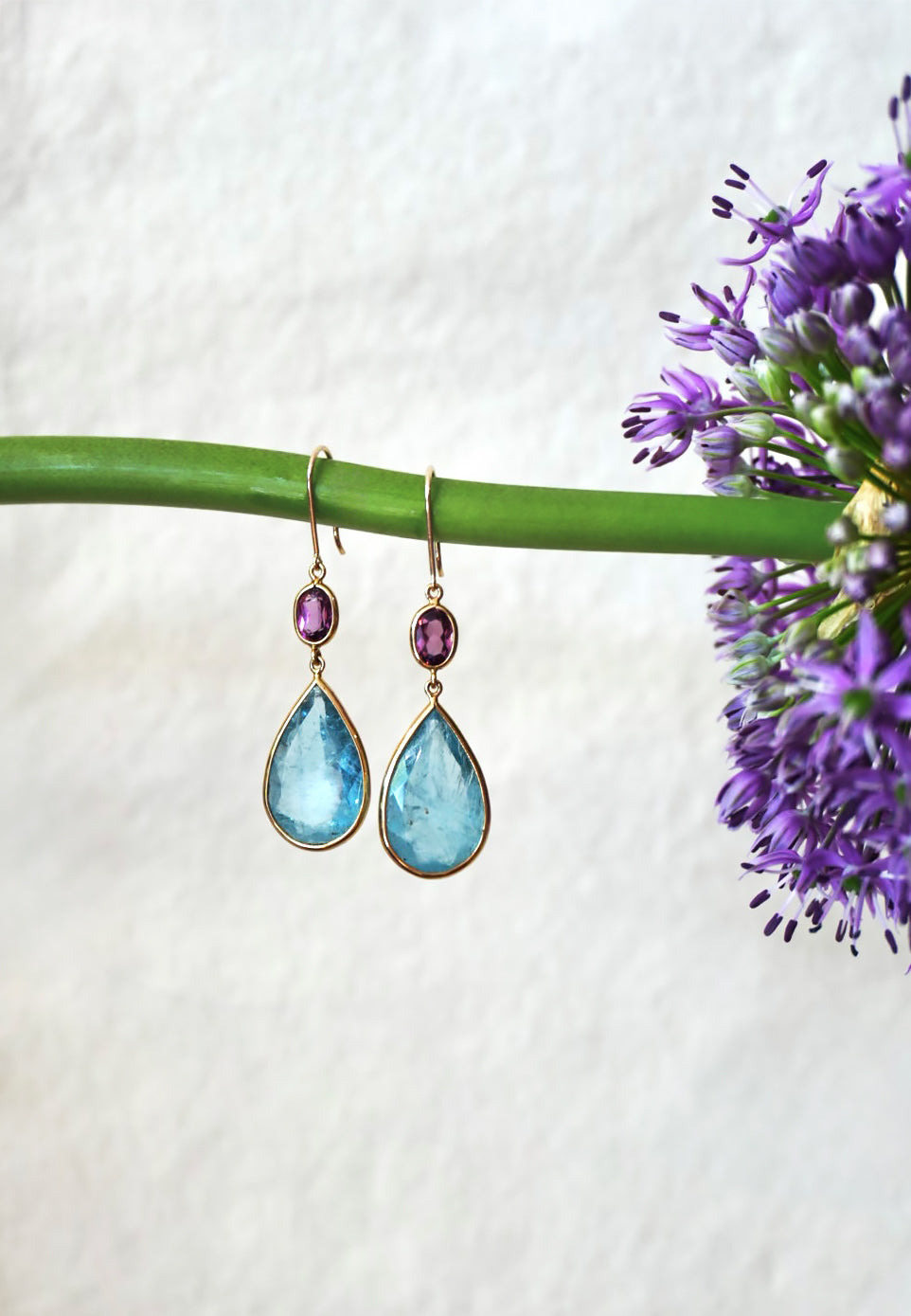 Teardrop-shaped earrings with aquamarine gemstones on a white background with purple flowers.