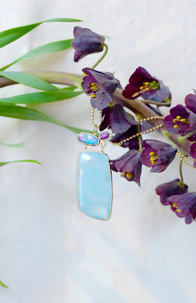 Large, light blue organic pendant necklace with gold chain on a white background with purple flowers