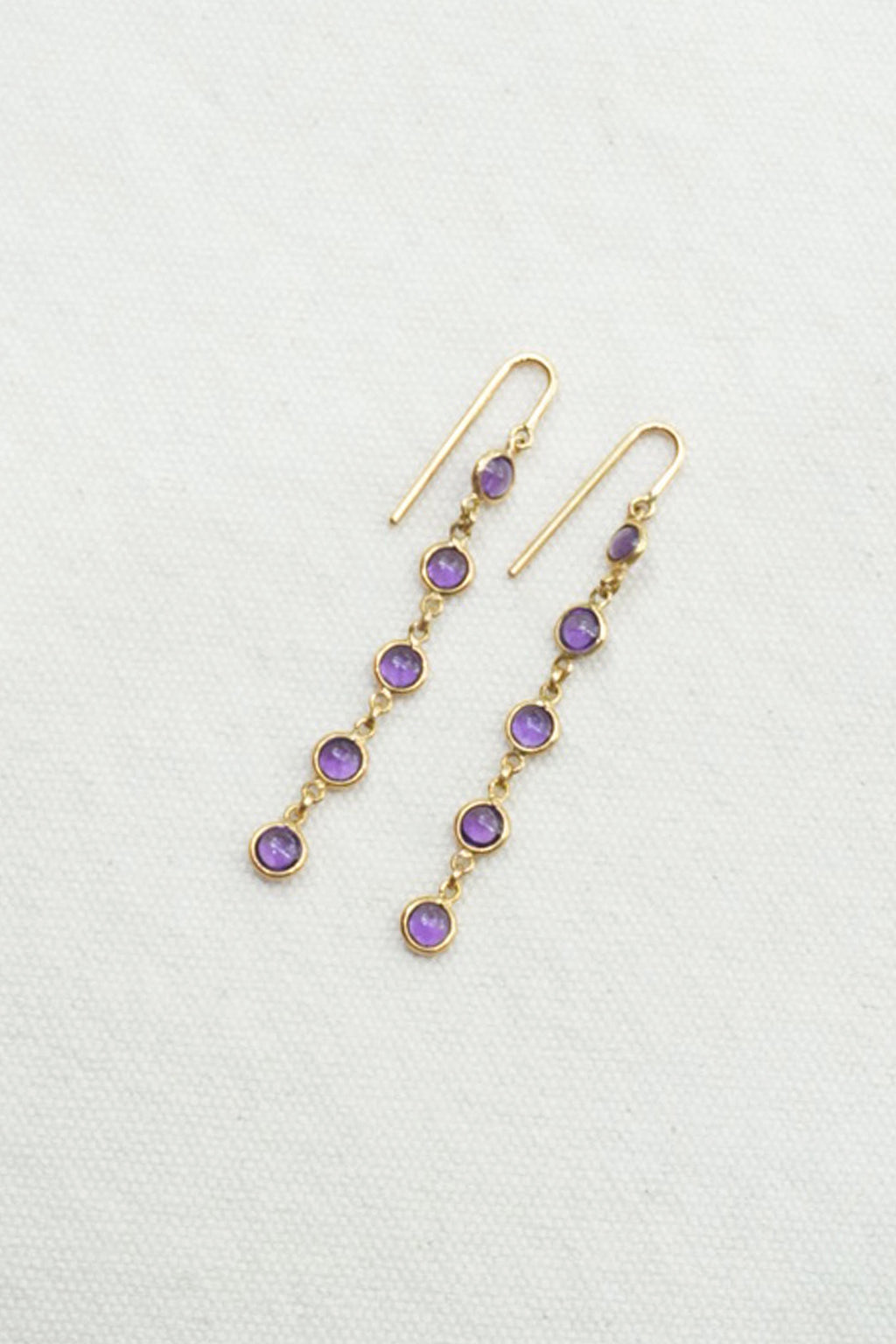 A pair of gold dangle earrings with small round purple gemstones, displayed on a textured off-white background.