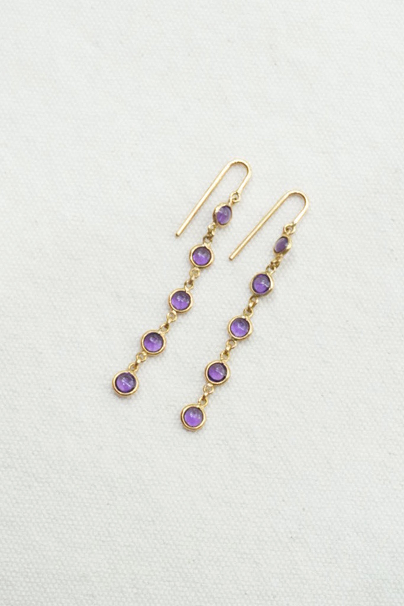 A pair of gold dangle earrings with small round purple gemstones, displayed on a textured off-white background.