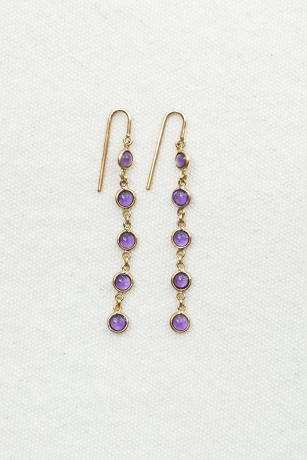 A pair of gold dangle earrings with five purple gemstones each, displayed on a textured off-white background.