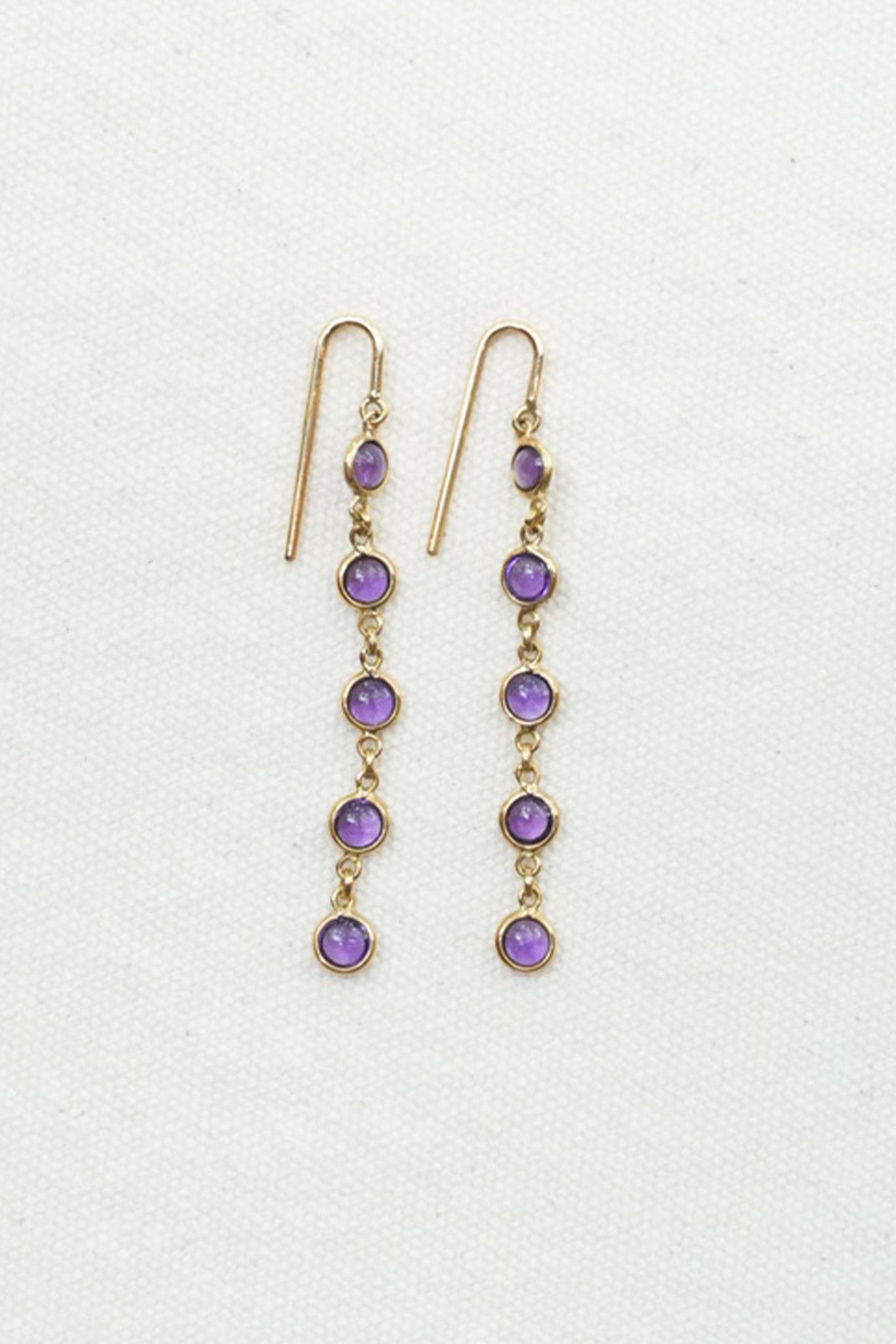 A pair of gold dangle earrings with five purple gemstones each, displayed on a textured off-white background.