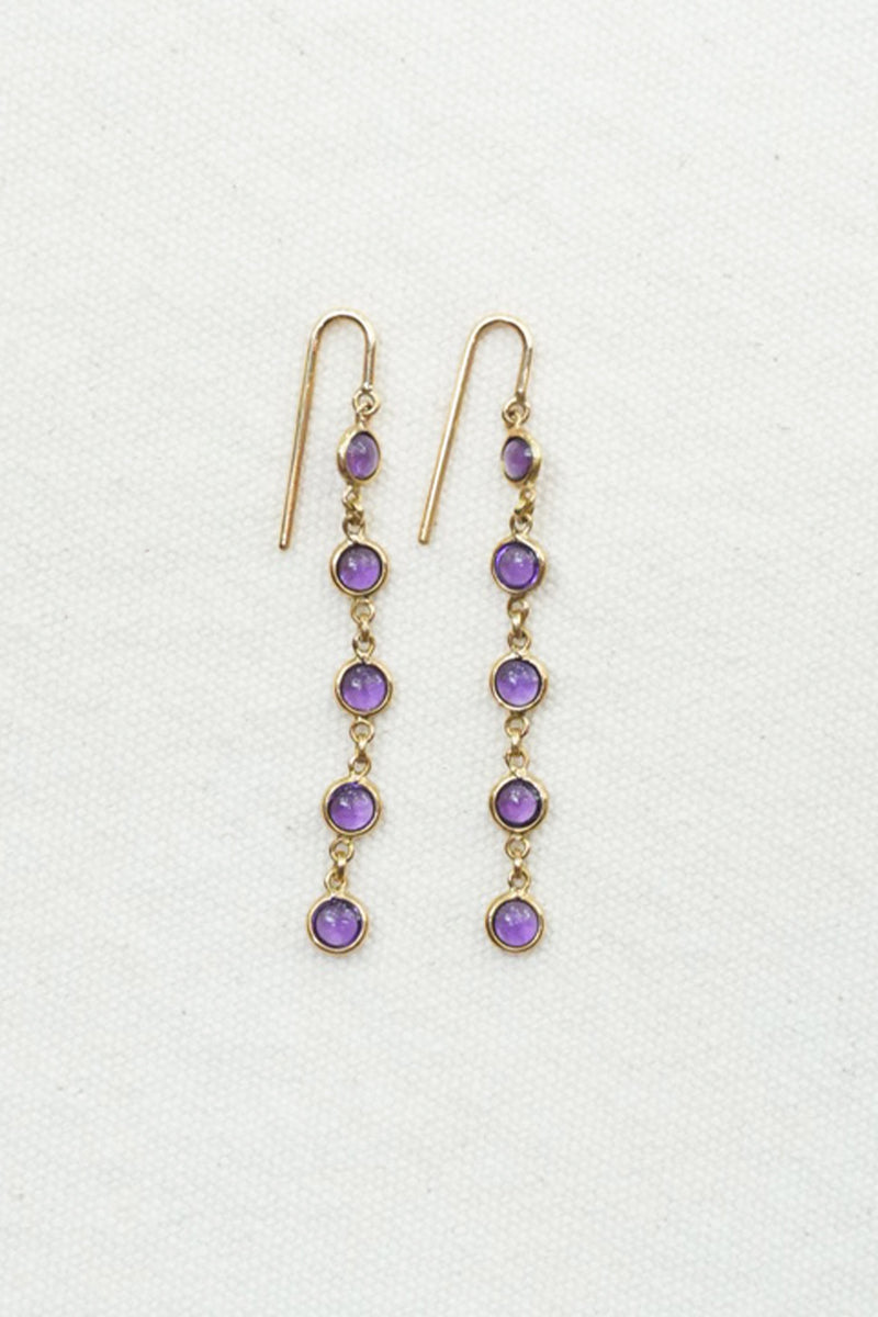 A pair of gold dangle earrings with five purple gemstones each, displayed on a textured off-white background.