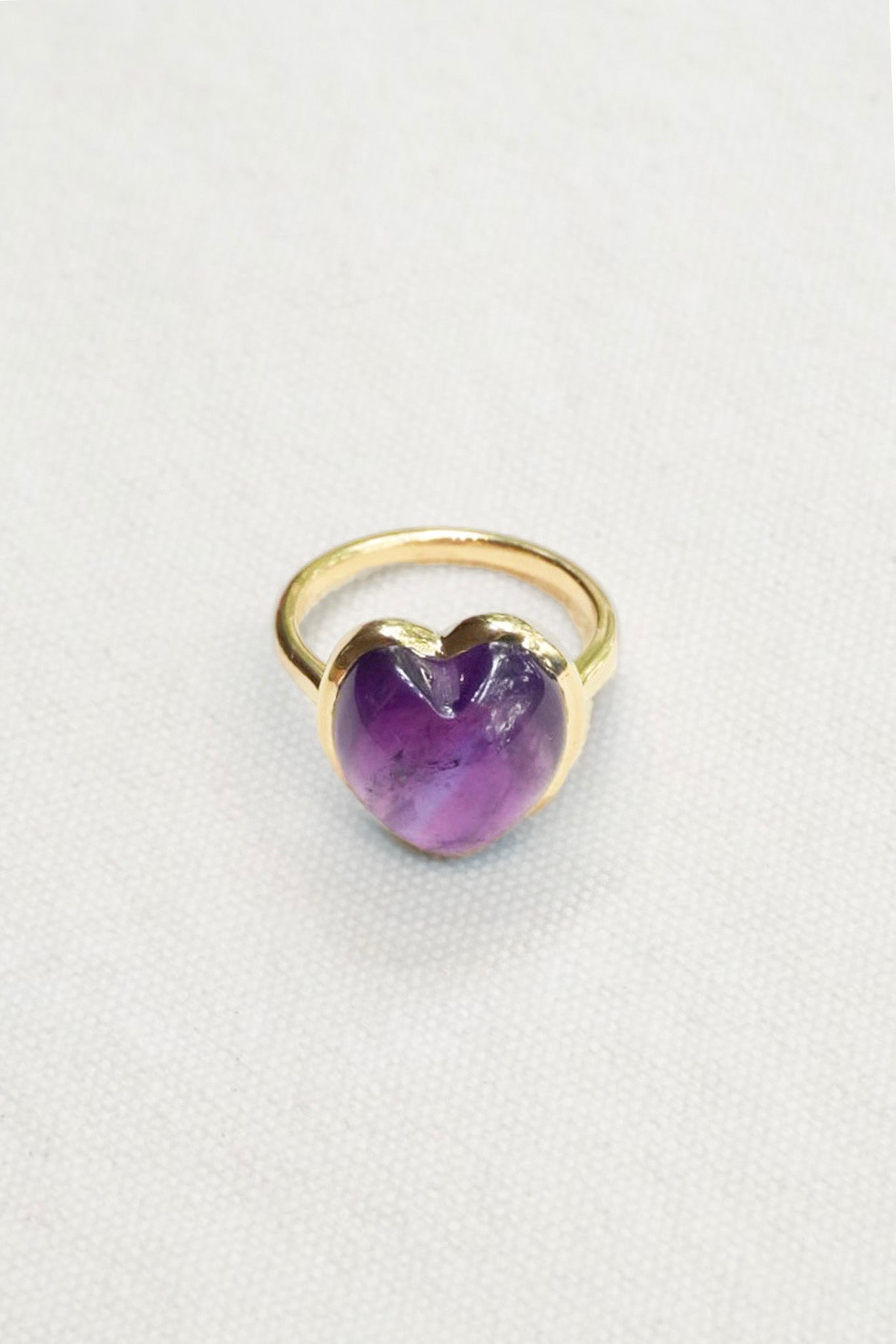 A gold ring featuring a heart-shaped purple gemstone, photographed on a textured off-white background.