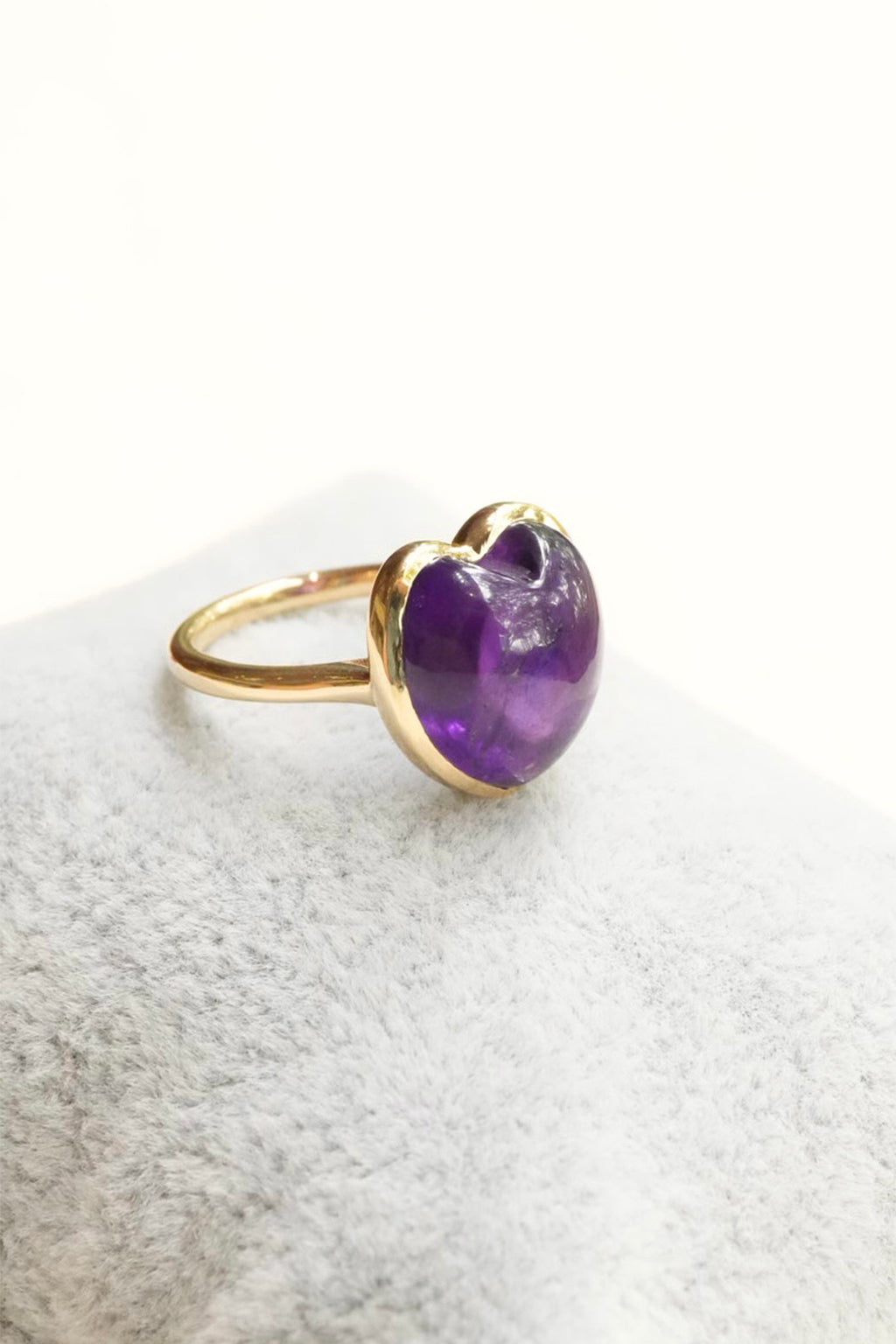 A gold ring with a heart-shaped purple amethyst gemstone, resting on a textured white surface.