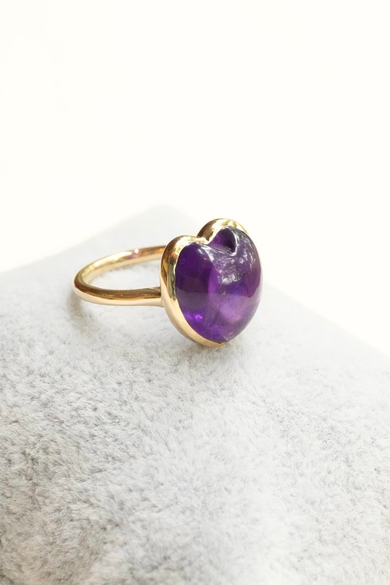 A gold ring with a heart-shaped purple amethyst gemstone, resting on a textured white surface.
