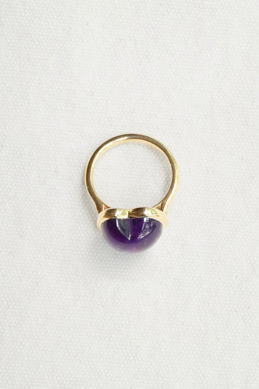 A gold ring featuring a polished, dome-shaped purple gemstone, photographed on a textured white fabric background.