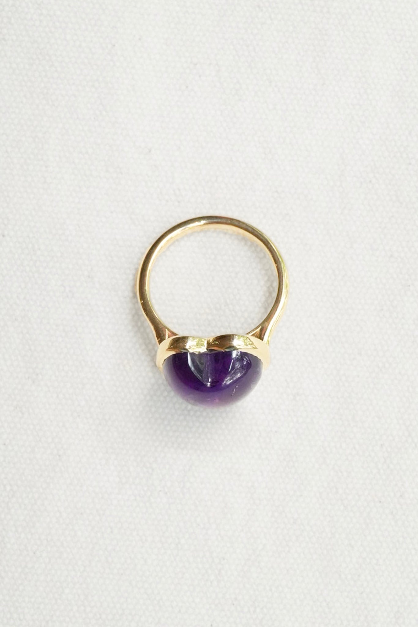 A gold ring featuring a polished, dome-shaped purple gemstone, photographed on a textured white fabric background.