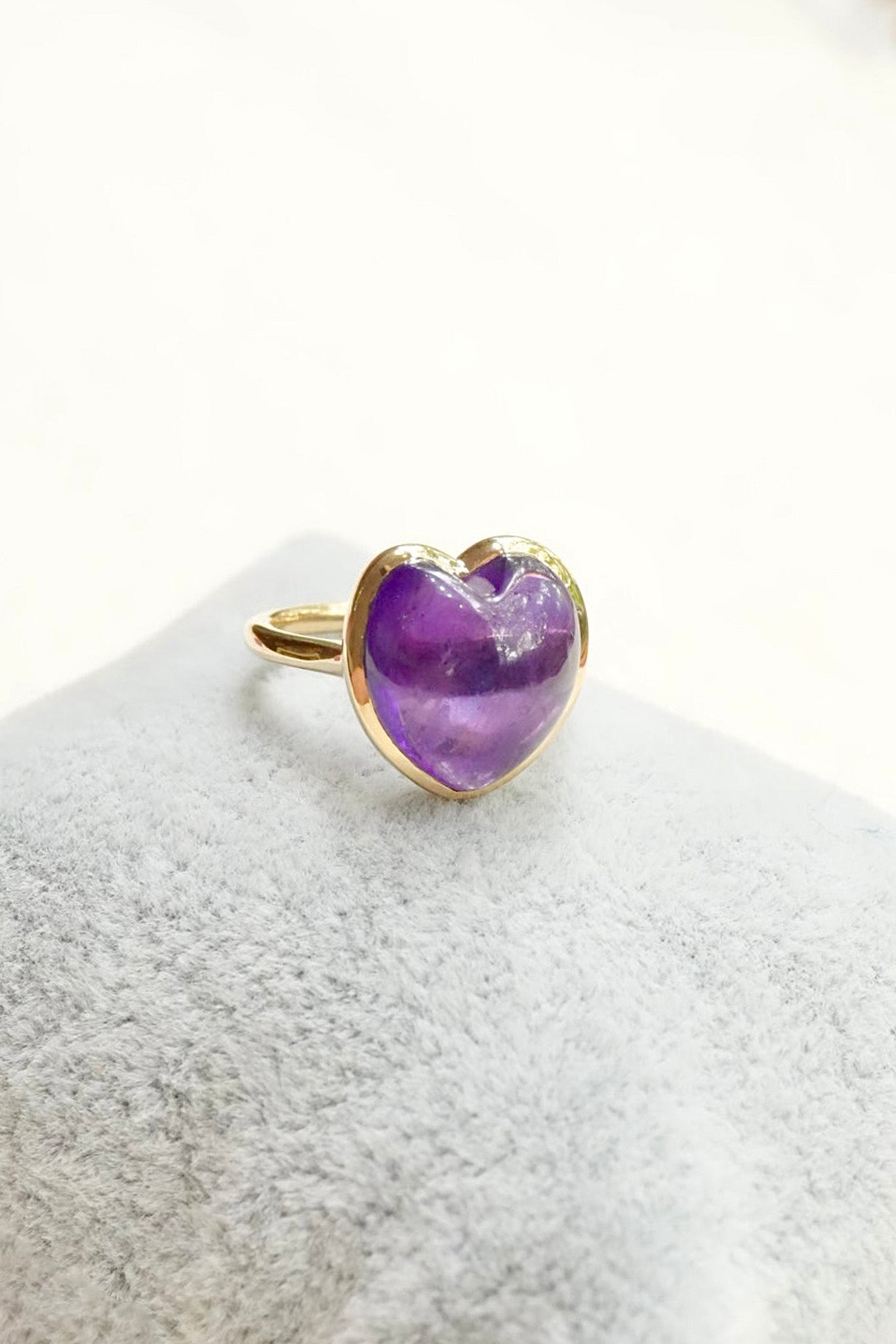 A gold heart-shaped ring with a purple amethyst center, displayed on a textured light gray surface against a white background.
