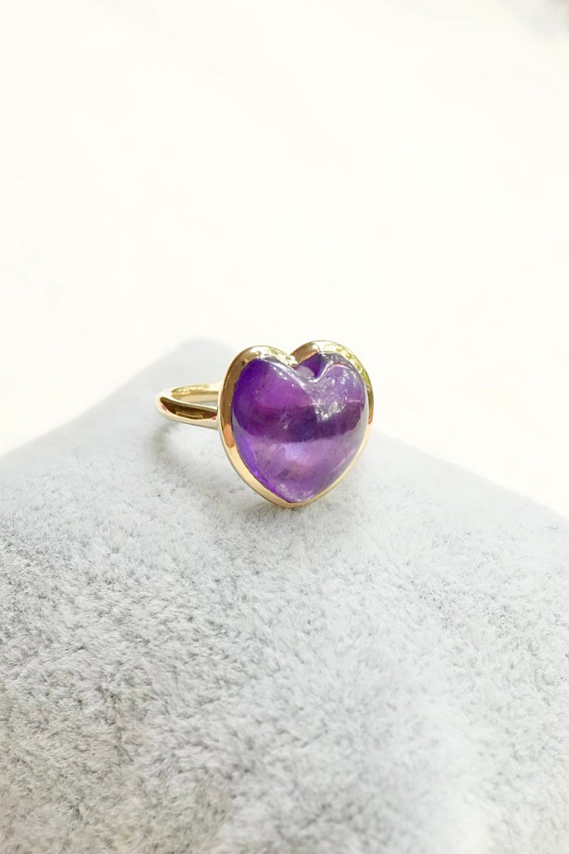 A gold heart-shaped ring with a purple amethyst center, displayed on a textured light gray surface against a white background.