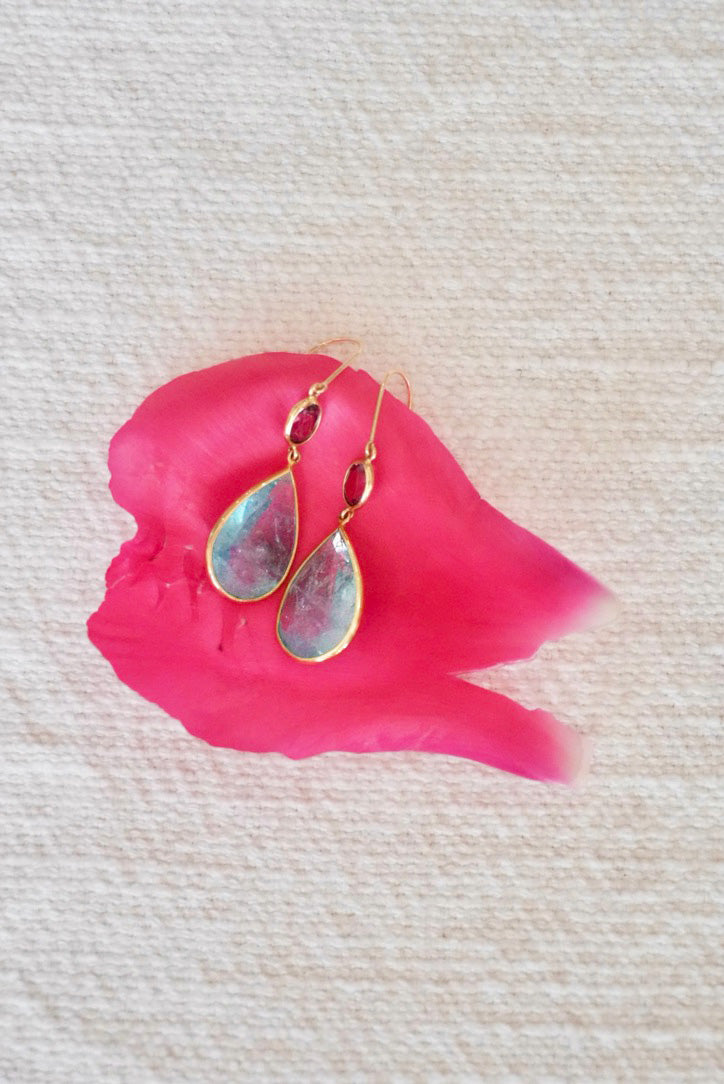Gold earrings with colorful aquamarine stones on a pink leaf against a textured beige background