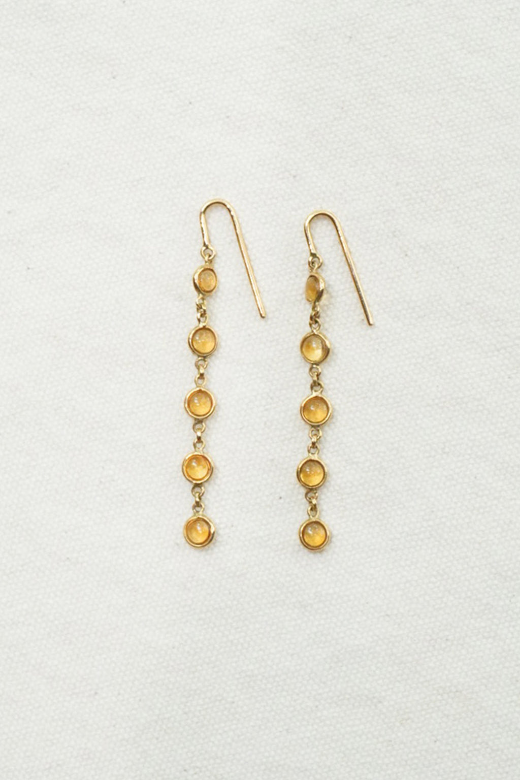 A pair of gold dangle earrings with five small round amber-yellow stones in each earring, displayed on a textured off-white background.