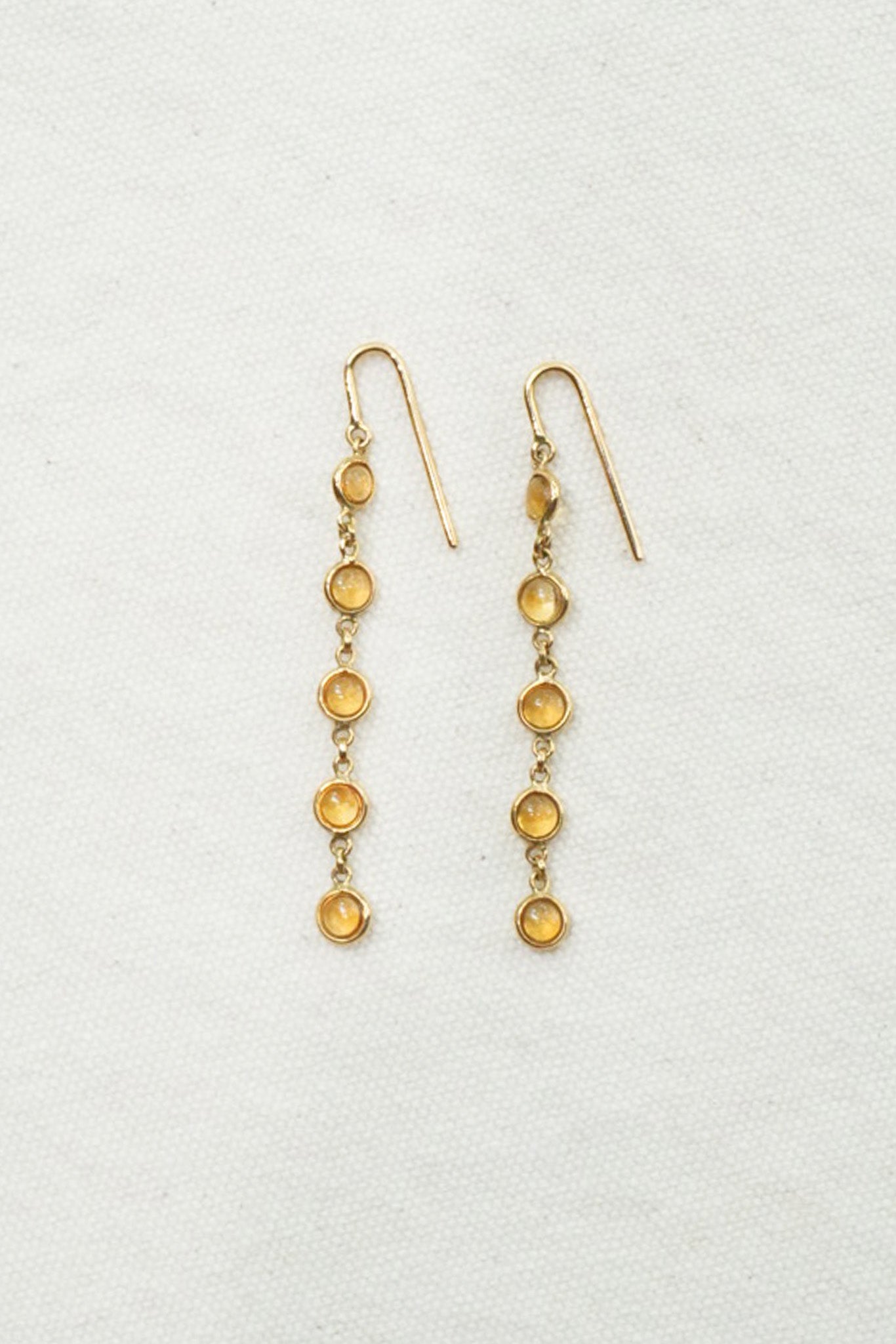 A pair of gold dangle earrings with five small round amber-yellow stones in each earring, displayed on a textured off-white background.