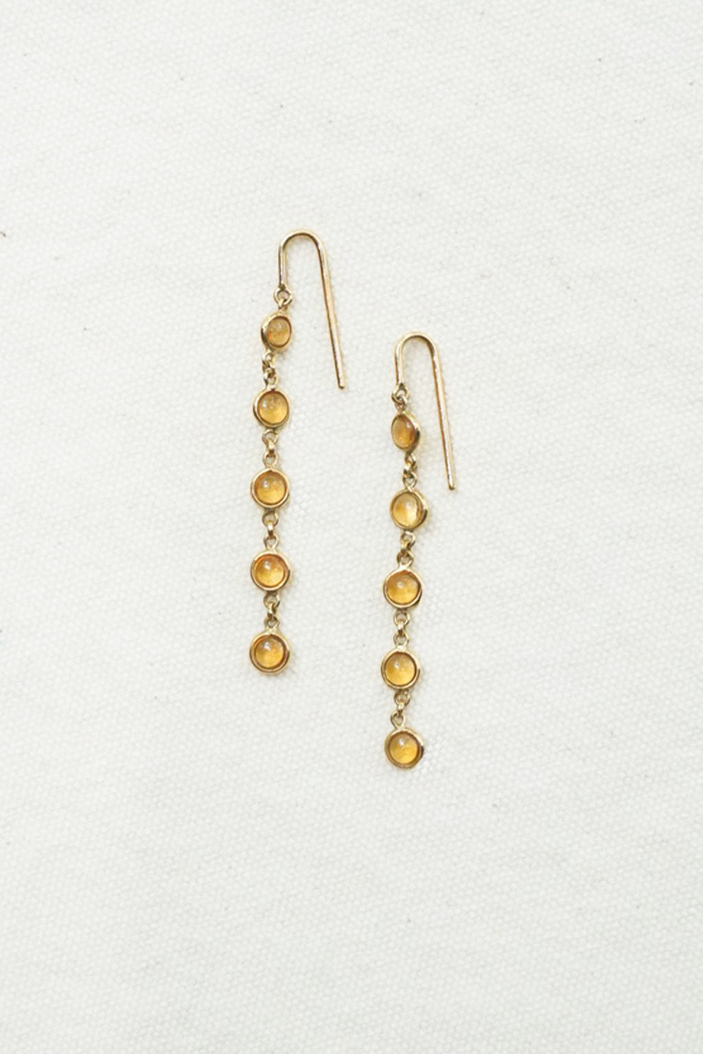 A pair of gold dangle earrings with small round amber-yellow beads, displayed on a textured off-white background.