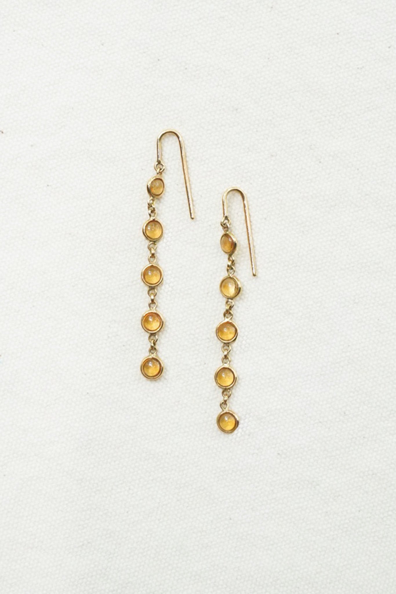 A pair of gold dangle earrings with small round amber-yellow beads, displayed on a textured off-white background.