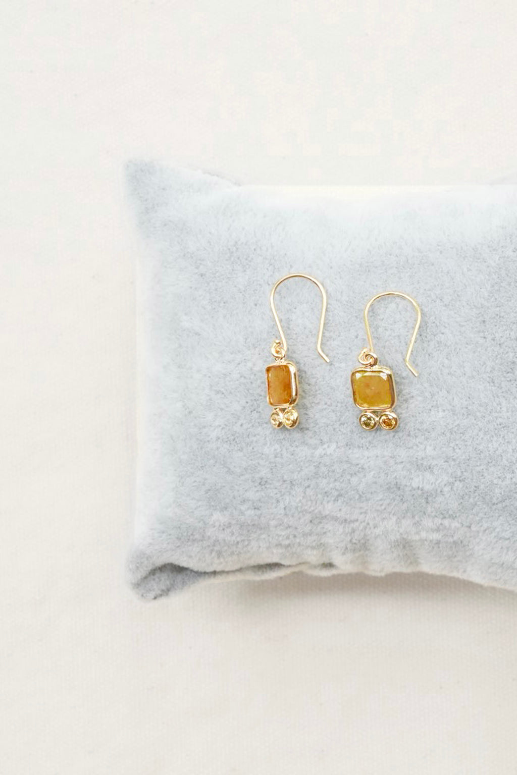 A pair of gold drop earrings with rectangular amber-yellow stones and small gold bead accents, displayed on a soft light gray pillow against a light neutral background.