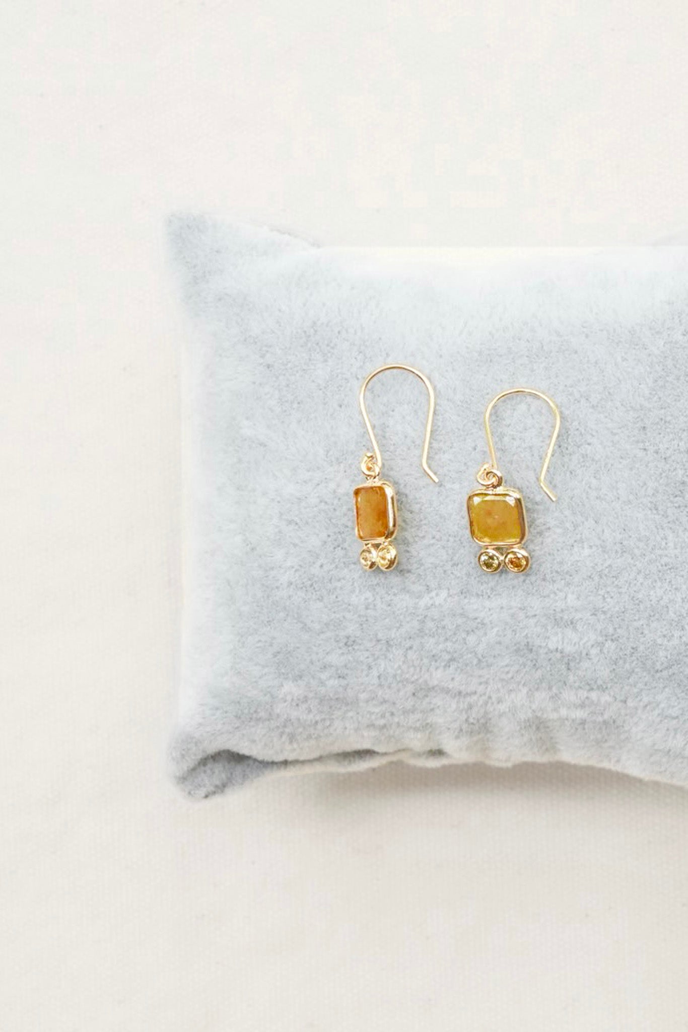 A pair of gold drop earrings with rectangular amber-yellow stones and small gold bead accents, displayed on a soft light gray pillow against a light neutral background.