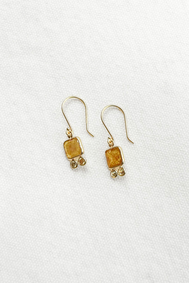 A pair of gold-toned dangle earrings with rectangular amber-yellow stone pendants and small bead accents, displayed on a textured white fabric background.