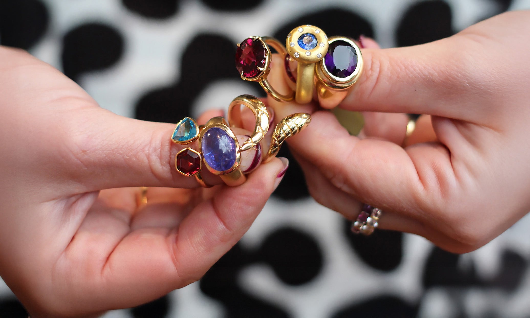 Two hands holding gold rings with colorful gemstones against a black and white patterned background.