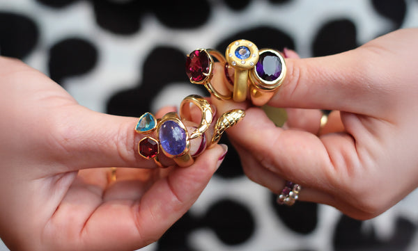 Two hands holding gold rings with colorful gemstones against a black and white patterned background.