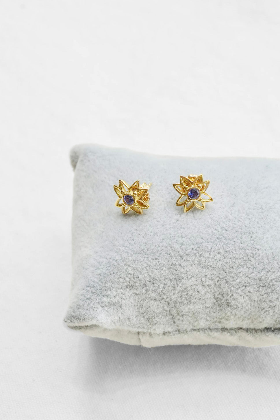 A pair of gold flower-shaped stud earrings with small dark blue gemstones in the center, displayed on a light gray velvet jewelry cushion against a white background.