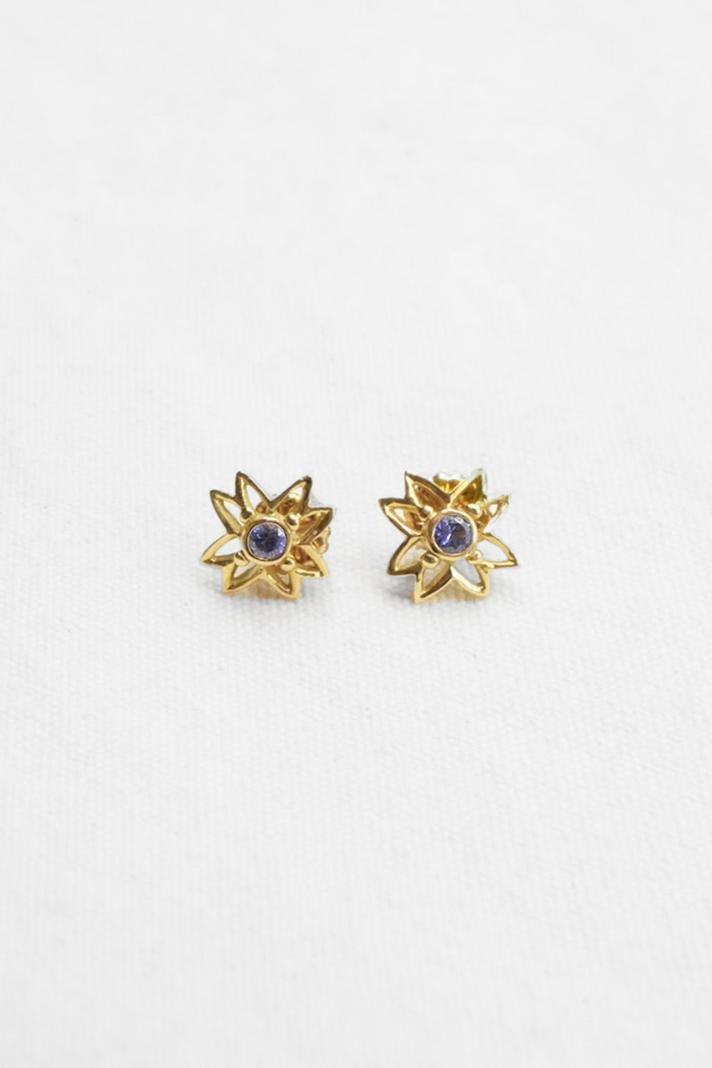 A pair of gold starburst stud earrings with a small round purple gemstone in the center, displayed on a clean white textured background.