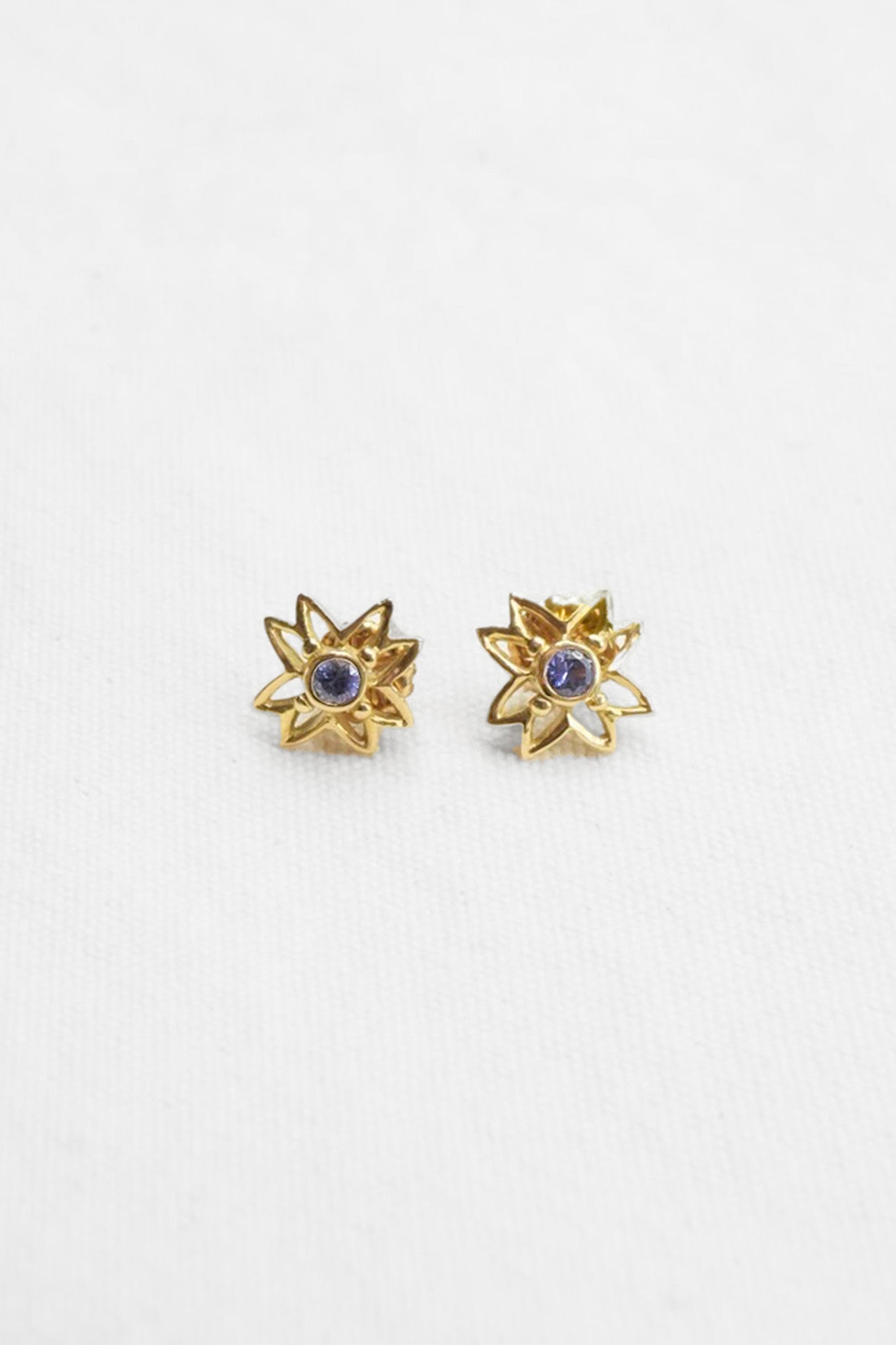 A pair of gold starburst stud earrings with a small round purple gemstone in the center, displayed on a clean white textured background.