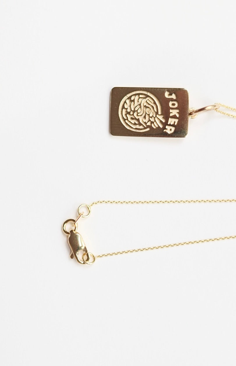 Gold necklace with a rectangular pendant featuring a circular design and 'Joker' text on a white background.