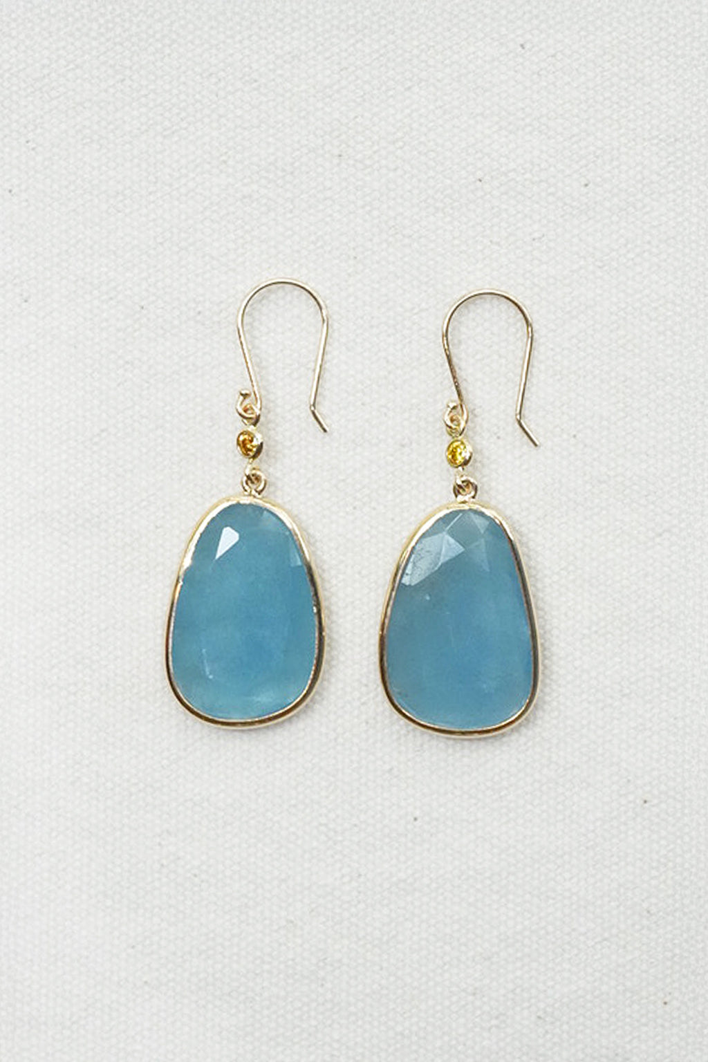 A pair of gold drop earrings with teardrop-shaped light blue gemstones, displayed on a textured white fabric background.