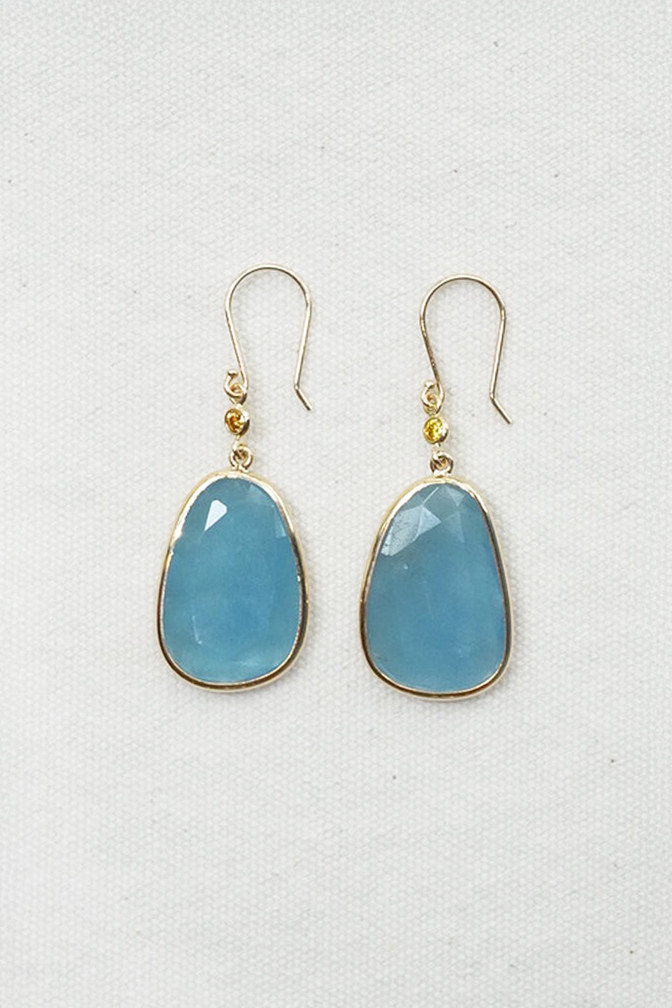 A pair of gold drop earrings with teardrop-shaped light blue gemstones, displayed on a textured white fabric background.