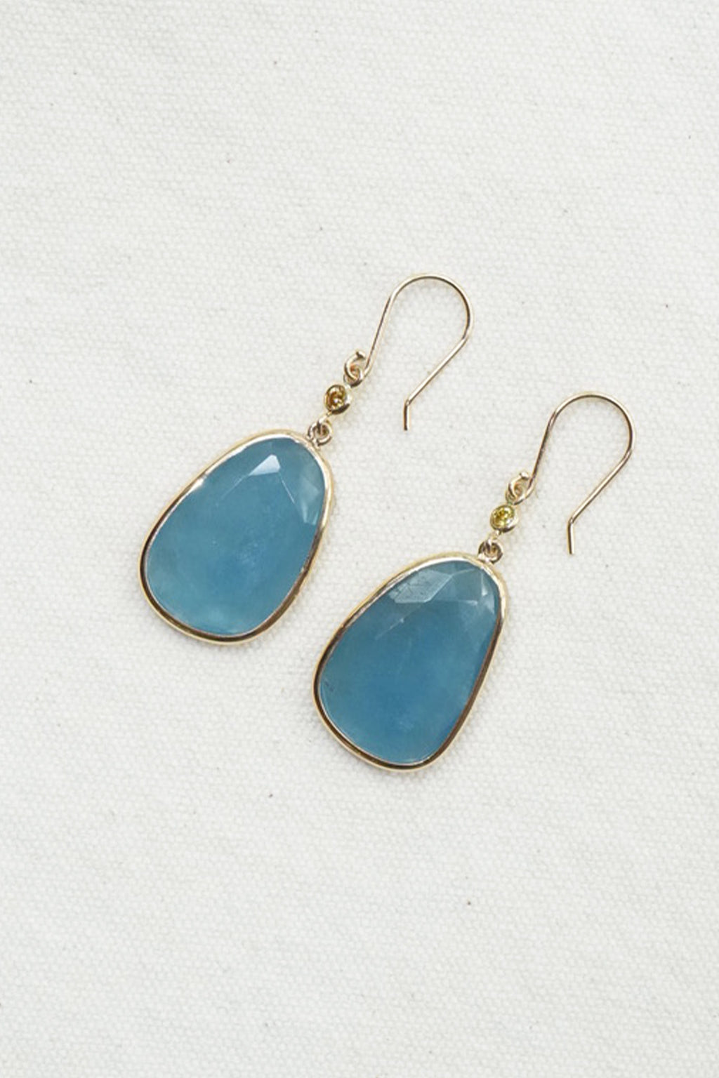 A pair of gold-tone drop earrings with smooth, teardrop-shaped light blue gemstones, displayed on a textured off-white background.