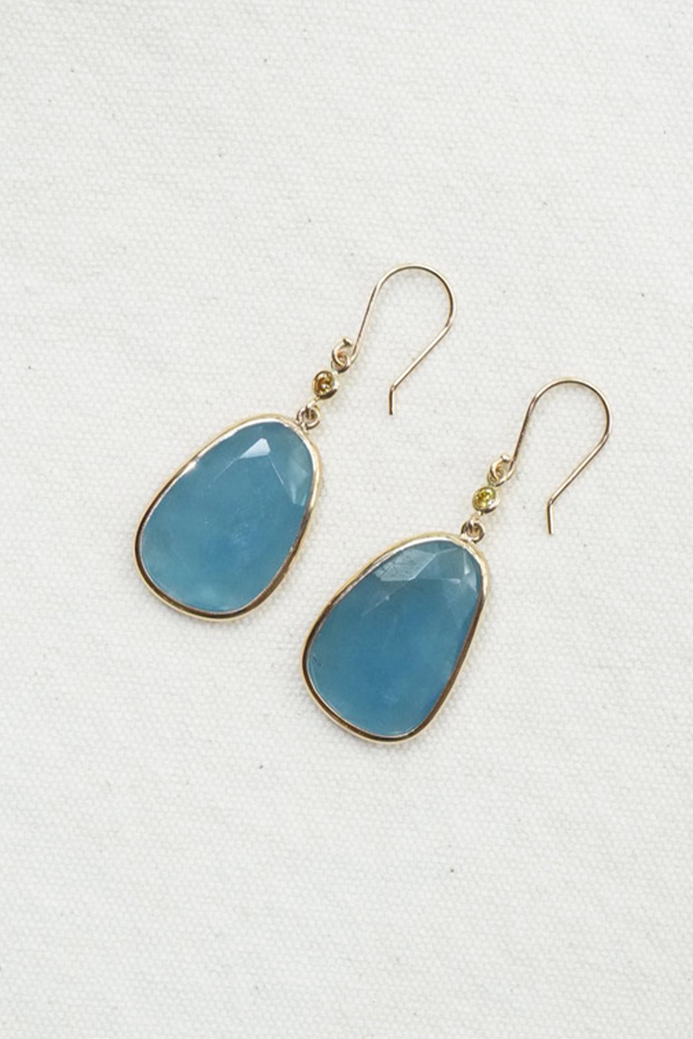 A pair of gold-tone drop earrings with smooth, teardrop-shaped light blue gemstones, displayed on a textured off-white background.