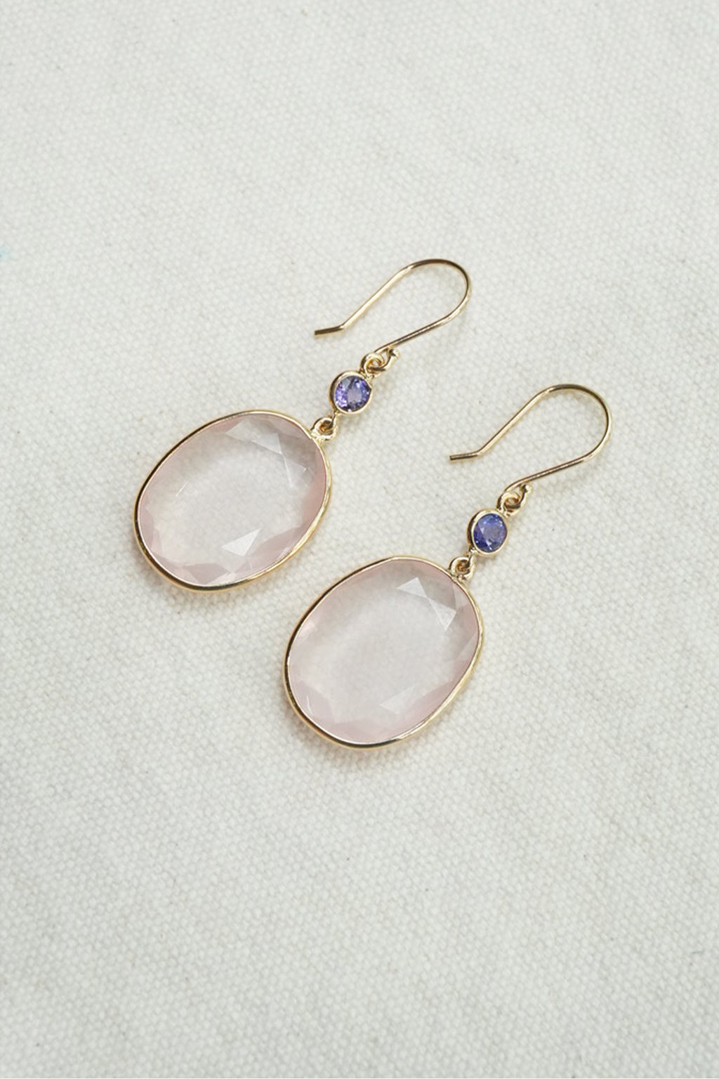 A pair of gold-tone drop earrings featuring oval pale pink stones and small round purple accent beads, displayed on a textured off-white background.