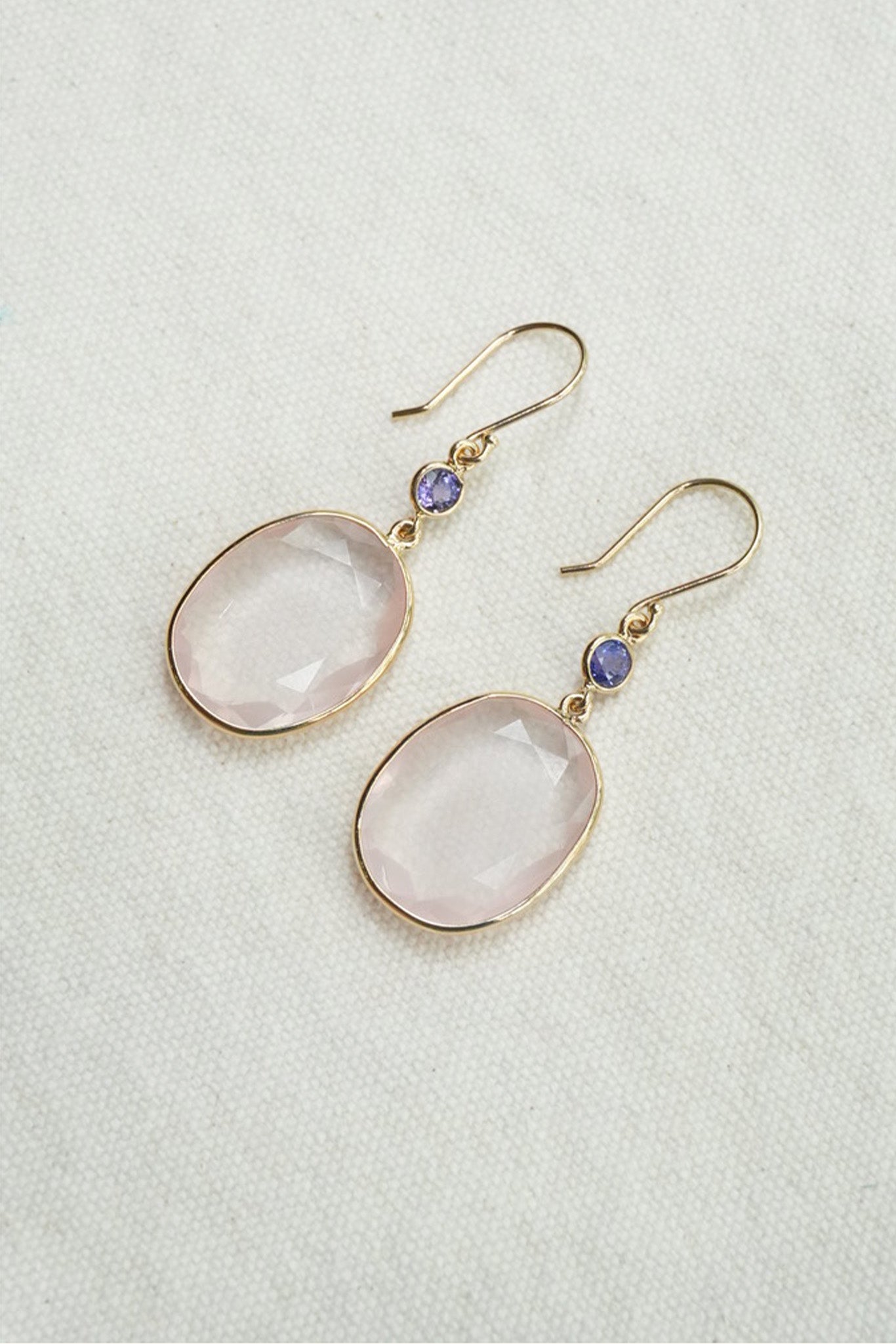 A pair of gold-tone drop earrings featuring oval pale pink stones and small round purple accent beads, displayed on a textured off-white background.