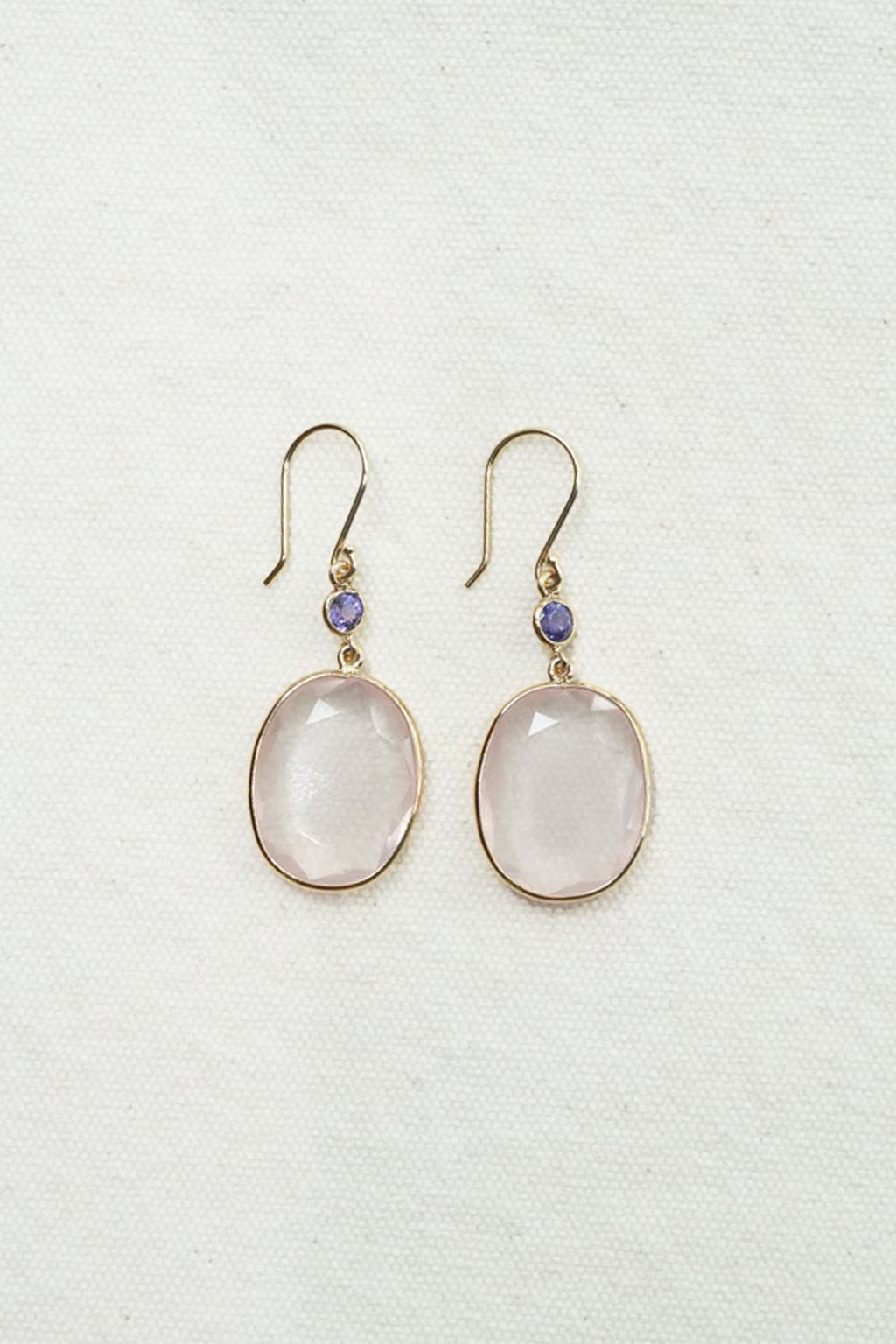 A pair of gold-tone drop earrings featuring oval pale pink stones and a small round purple accent bead above each stone, displayed on a textured off-white fabric background.