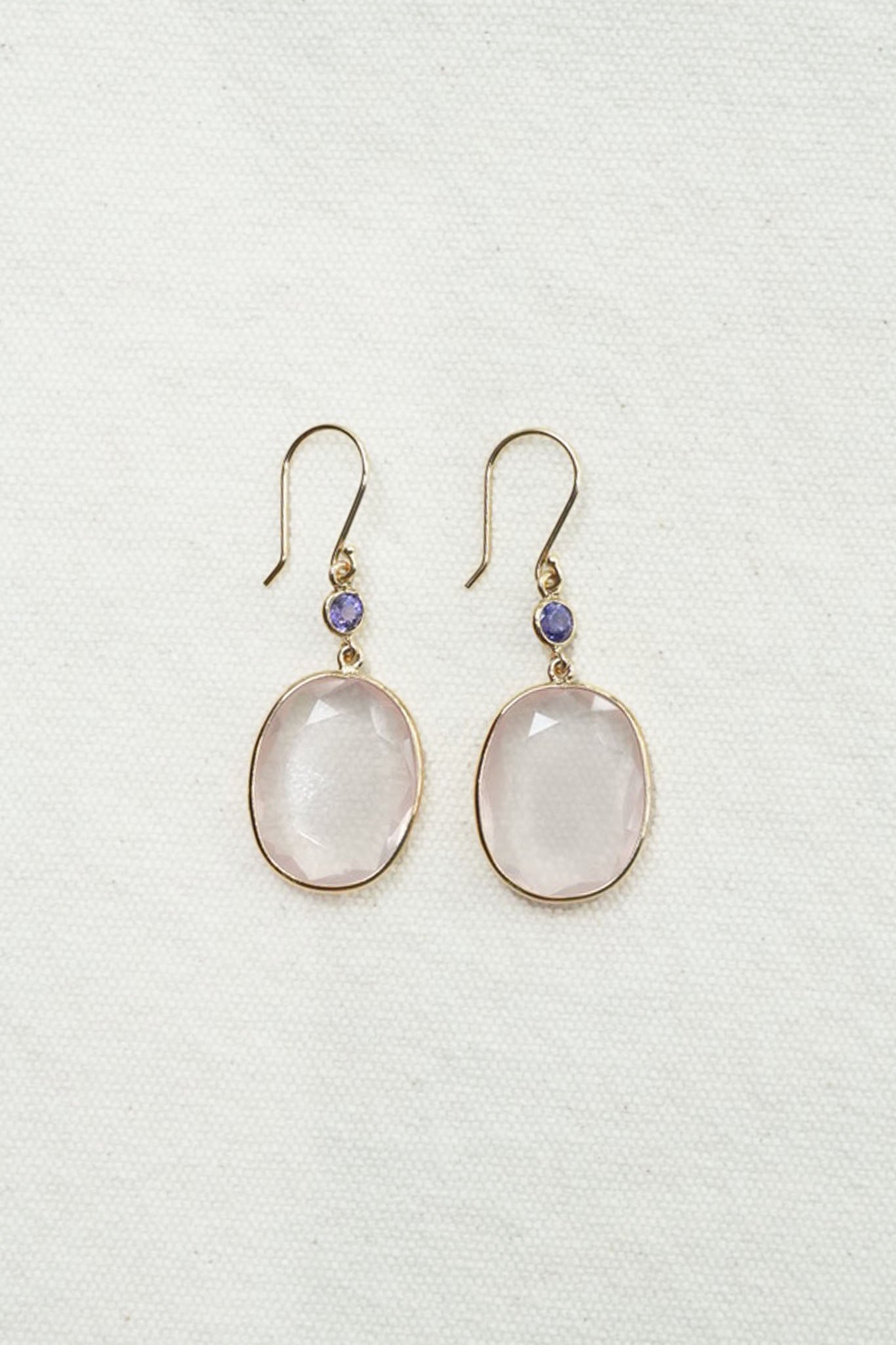 A pair of gold-tone drop earrings featuring oval pale pink stones and a small round purple accent bead above each stone, displayed on a textured off-white fabric background.