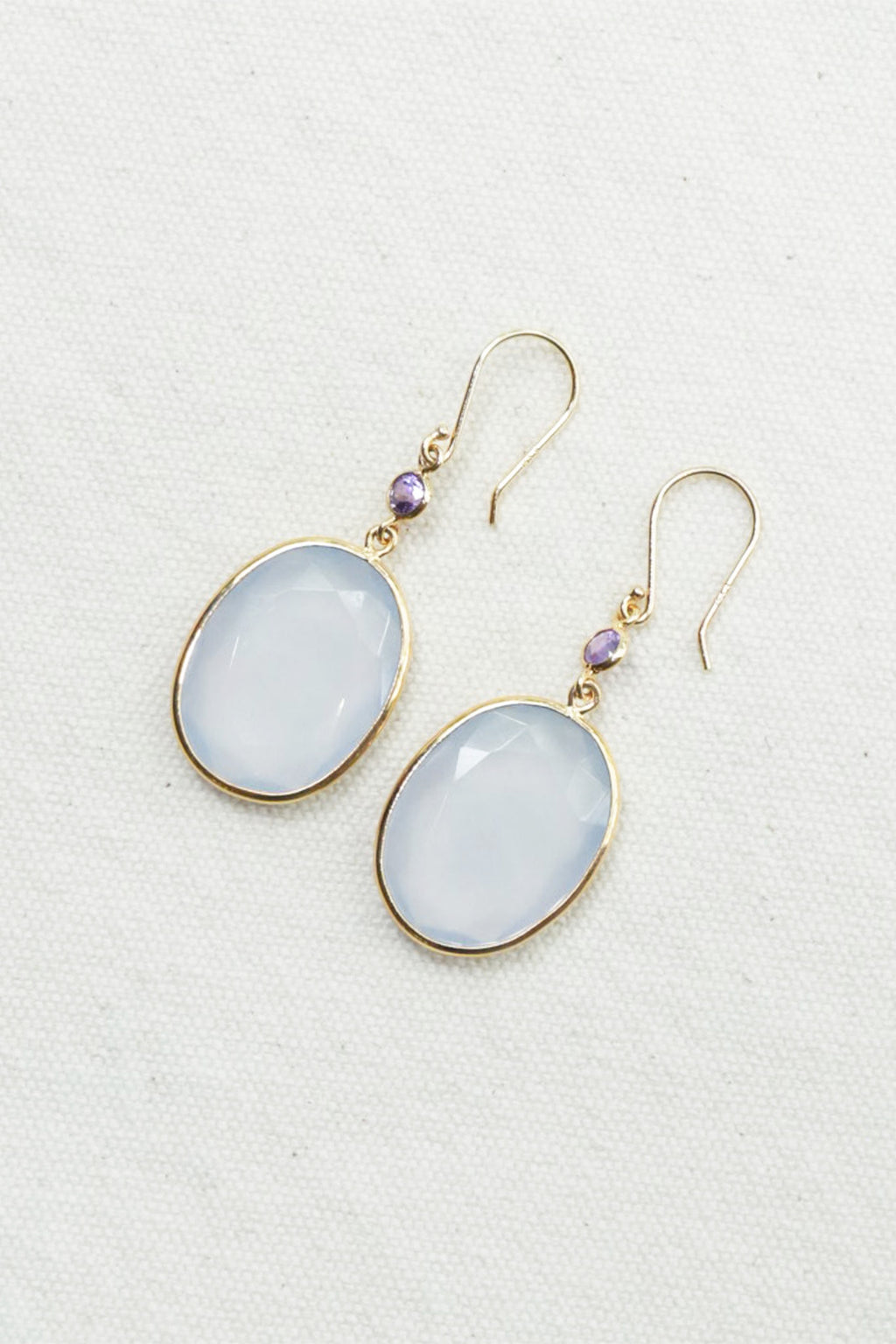 A pair of gold-tone drop earrings featuring oval milky white cabochon gemstones and small round purple accent beads, displayed on a textured off-white fabric background.