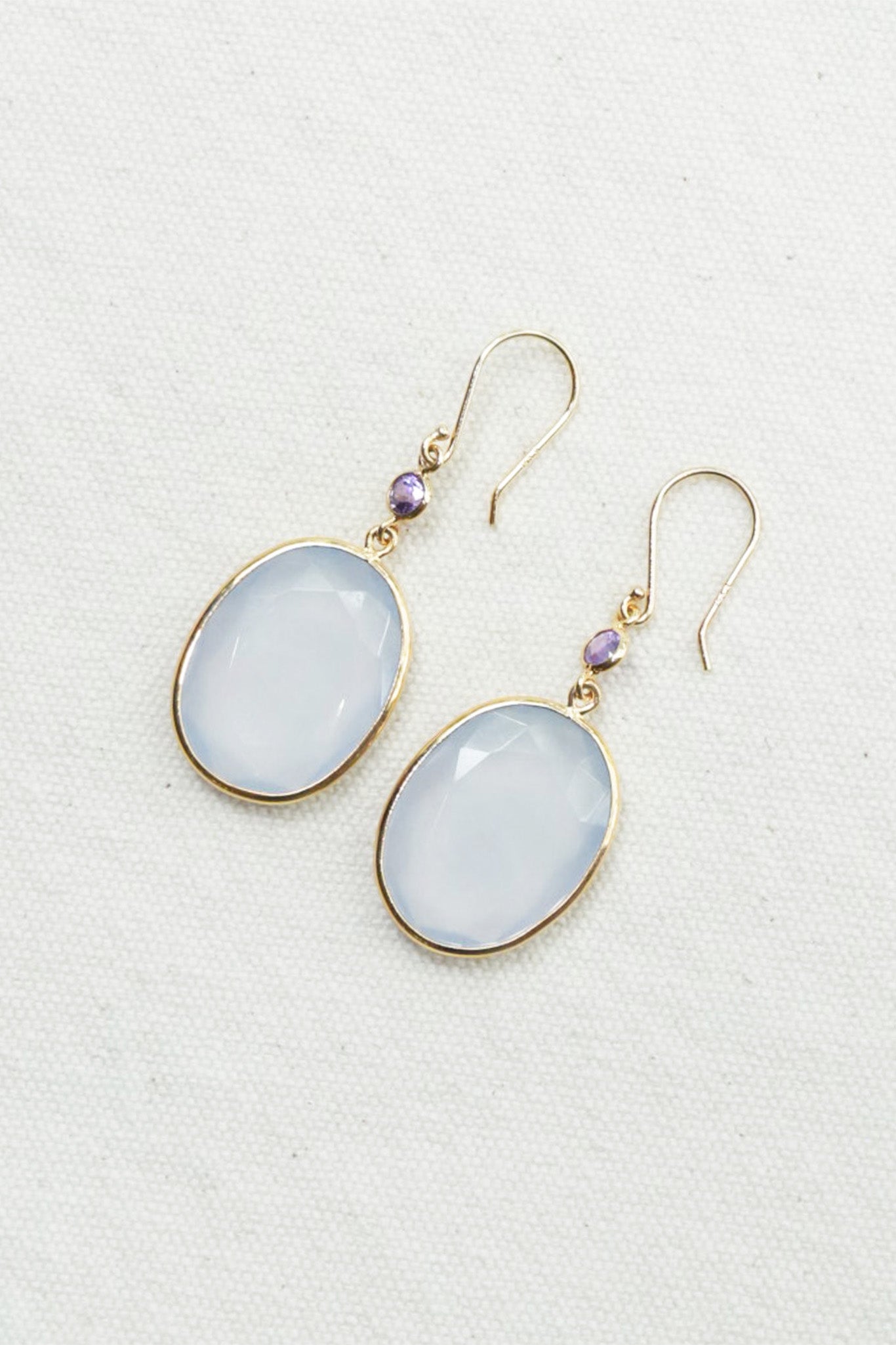 A pair of gold-tone drop earrings featuring oval milky white cabochon gemstones and small round purple accent beads, displayed on a textured off-white fabric background.