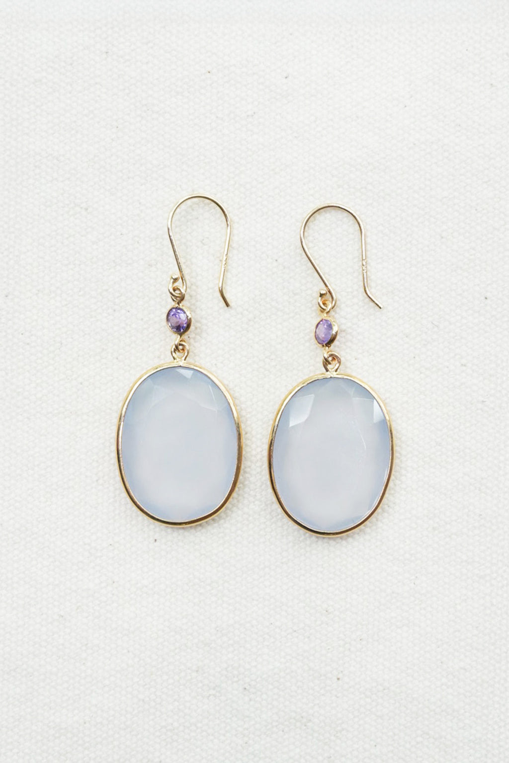 A pair of gold-tone drop earrings featuring oval milky white cabochon stones and small purple accent beads, displayed on a textured off-white fabric background.