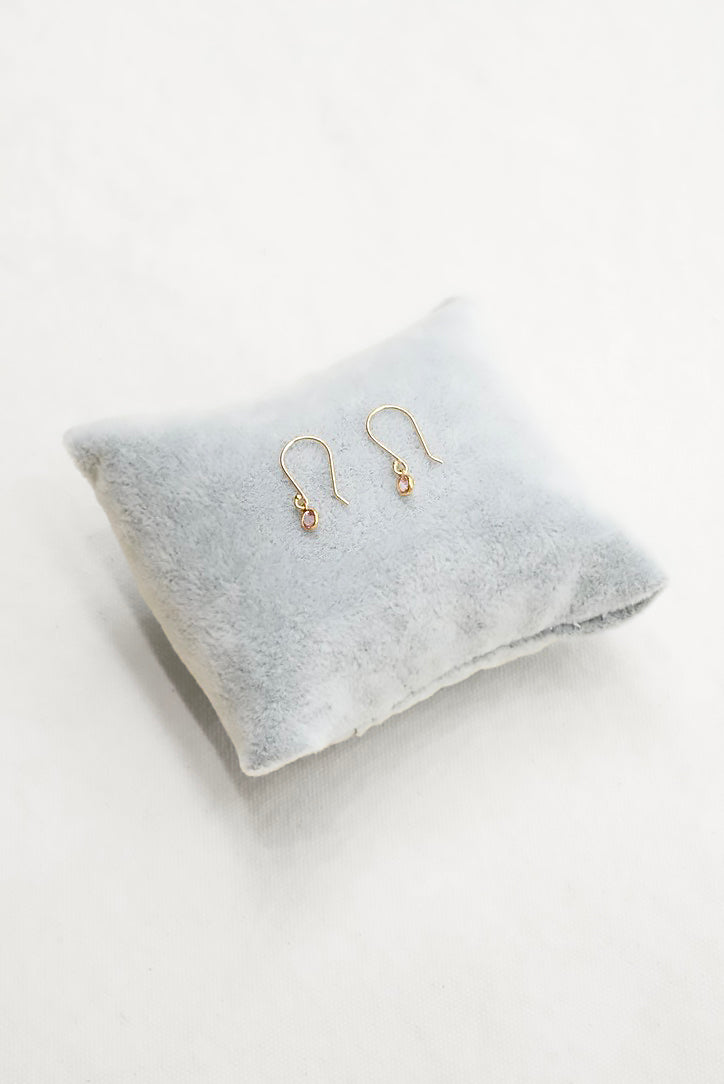 A pair of delicate gold dangle earrings resting on a small, light gray plush velvet pillow against a clean white background.