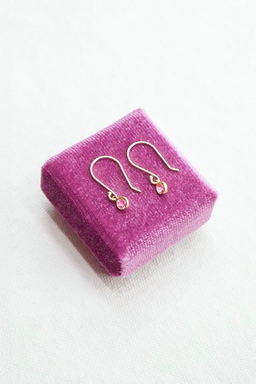 A pair of small gold-tone dangle earrings with tiny pink gemstones, displayed on a small textured purple velvet jewelry box against a white background.