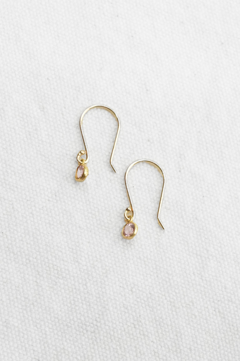 A pair of delicate gold drop earrings with small clear stones and pale pink teardrop-shaped gemstones, displayed on a textured white fabric background.