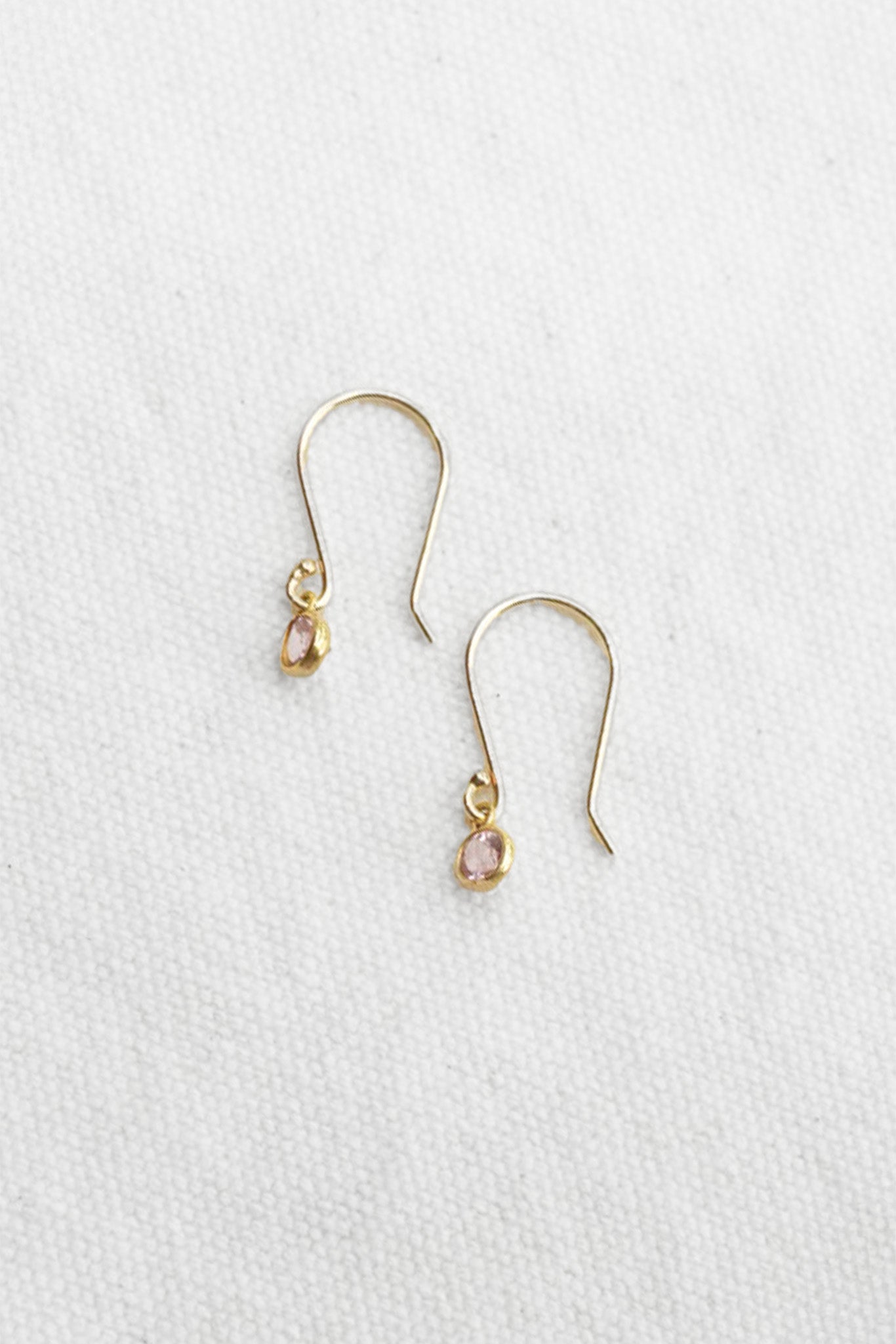 A pair of delicate gold drop earrings with small clear stones and pale pink teardrop-shaped gemstones, displayed on a textured white fabric background.