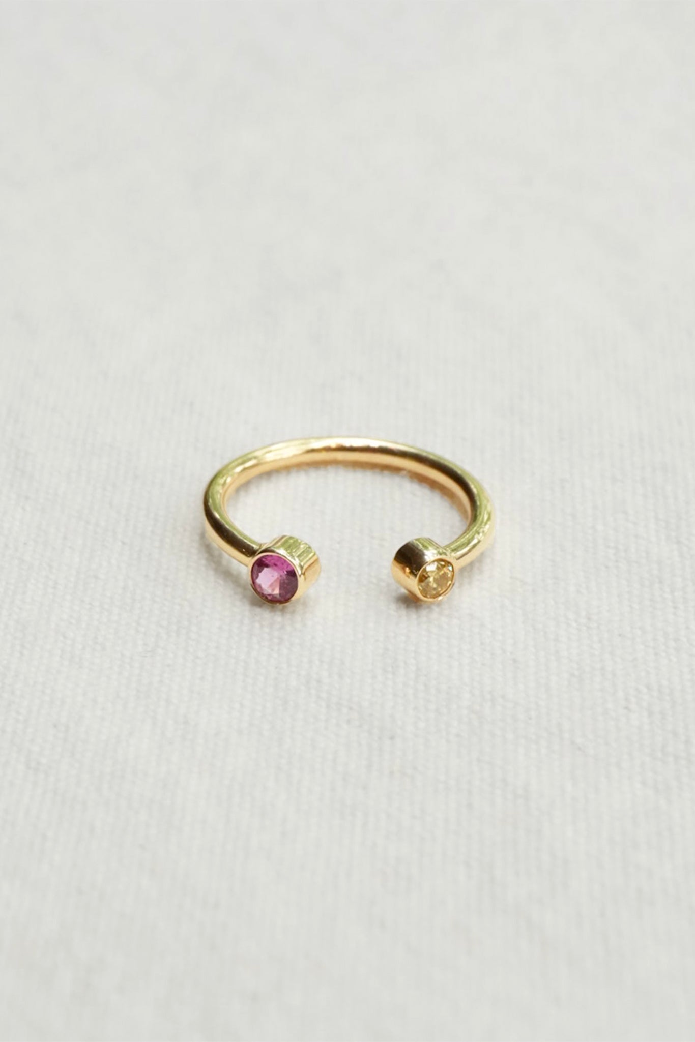 A delicate gold open-ended ring featuring a small round pink gemstone on one side and a yellow gemstone on the other, displayed on a textured off-white fabric background.