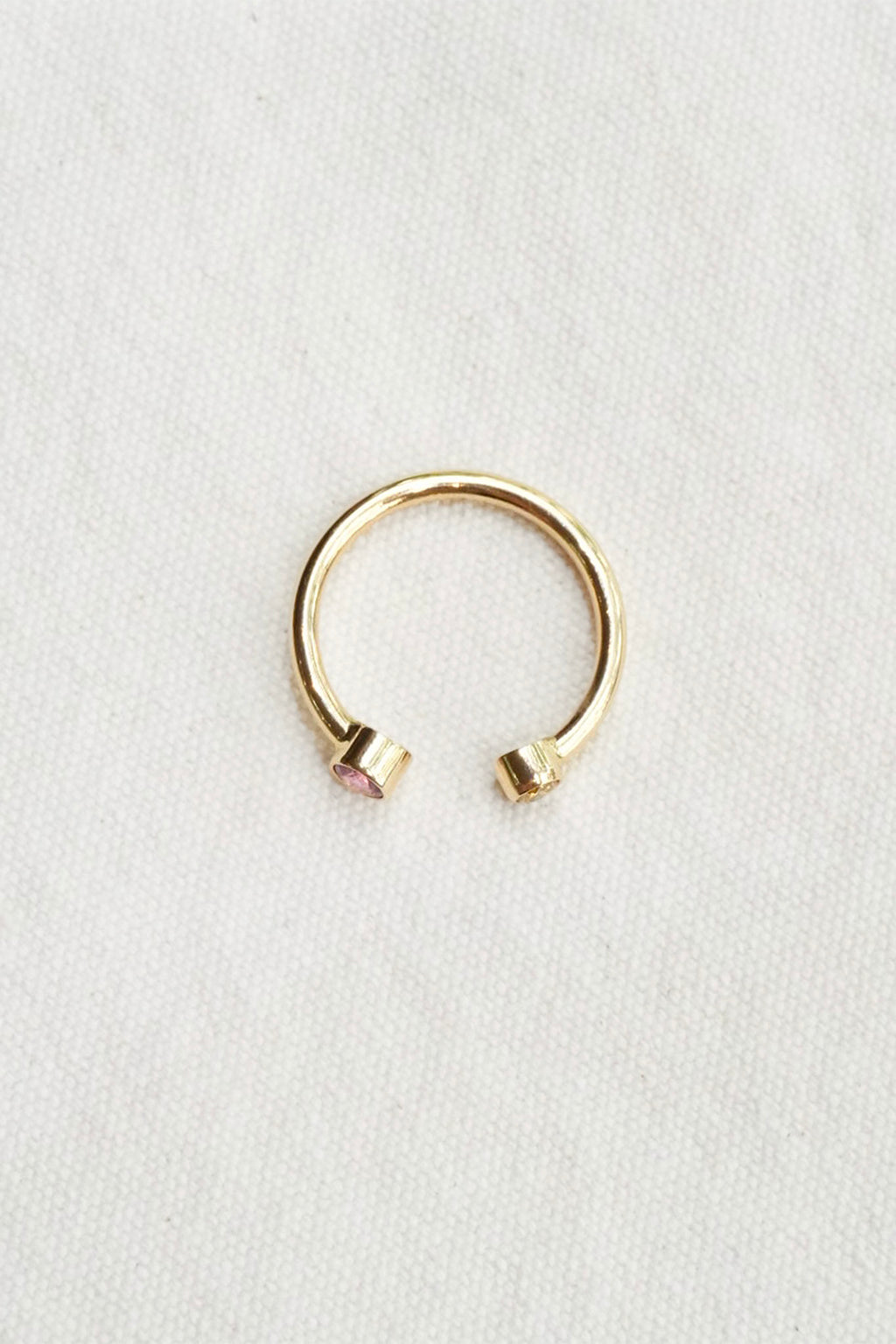 A delicate gold-toned open ring with small pink and yellow gemstones at each end, displayed on a textured white fabric background.