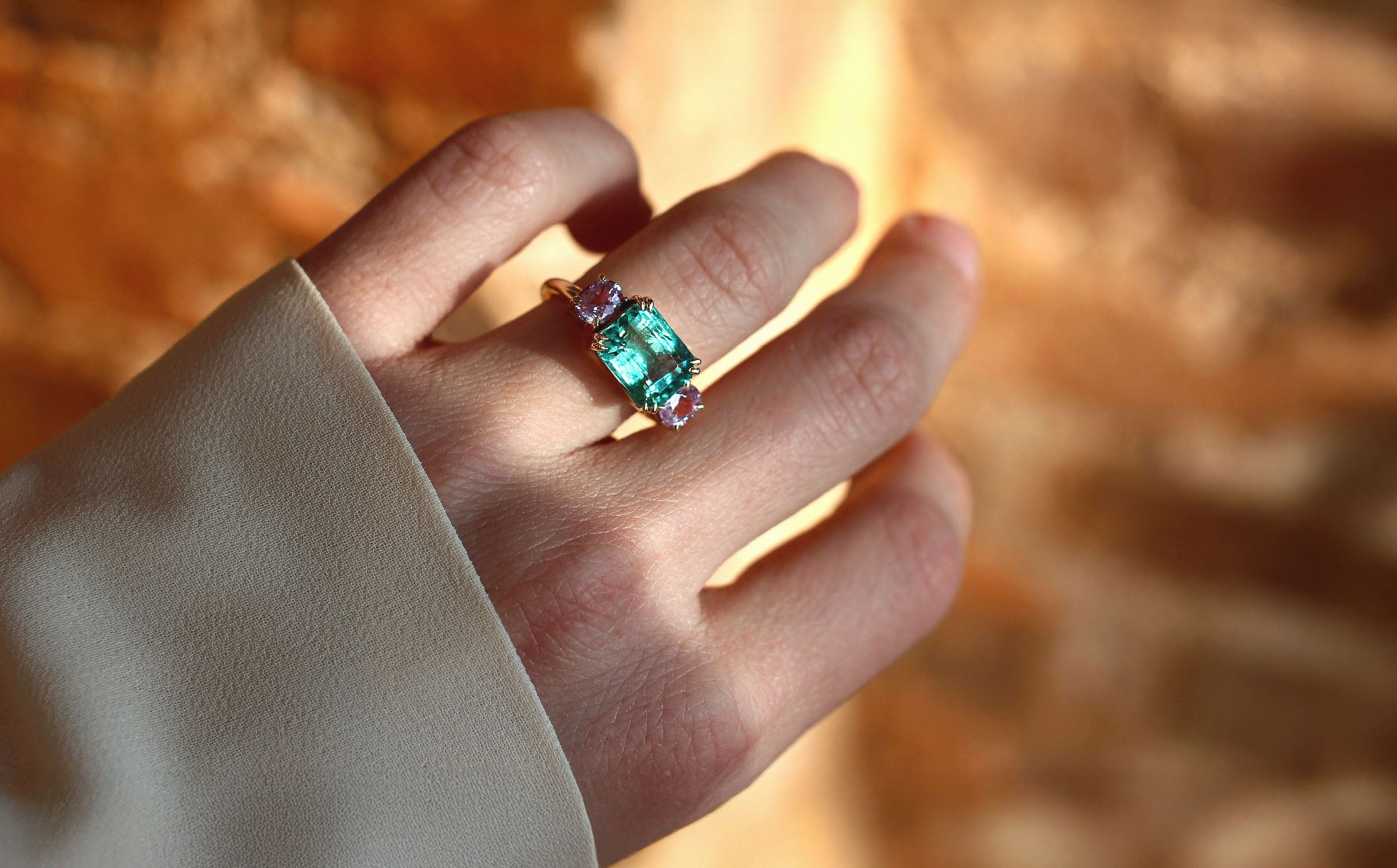 Hand wearing a green emerald ring with pink oval side gemstones against a blurred background