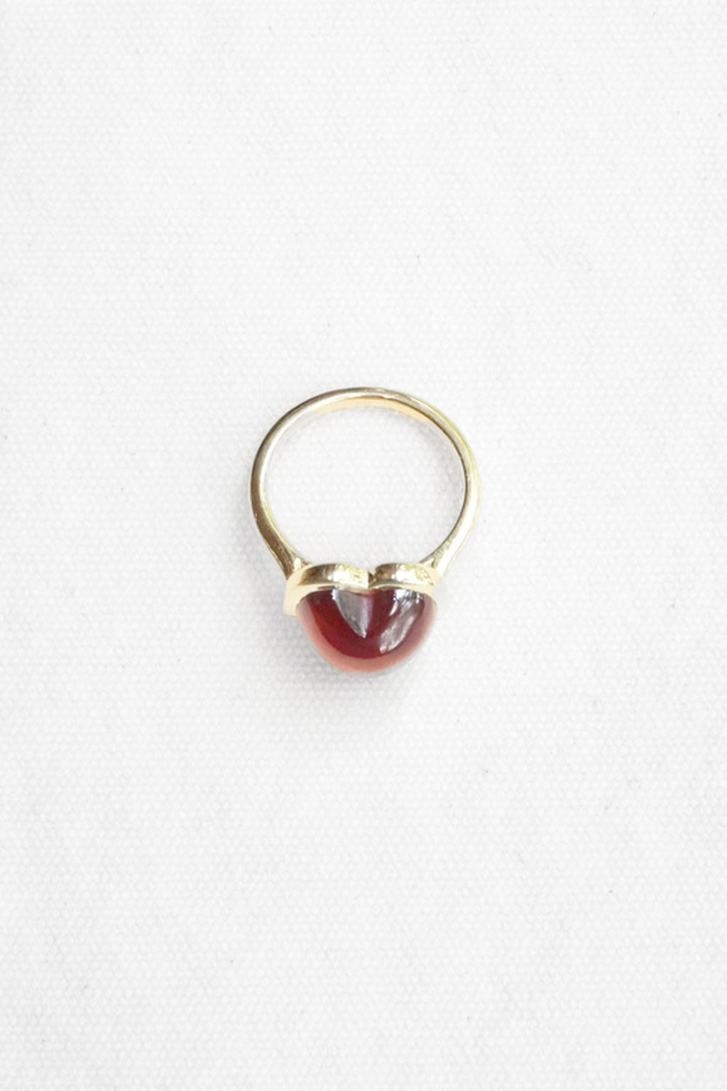 A gold ring featuring a small, polished red gemstone in a heart-shaped bezel setting, photographed on a clean white background.