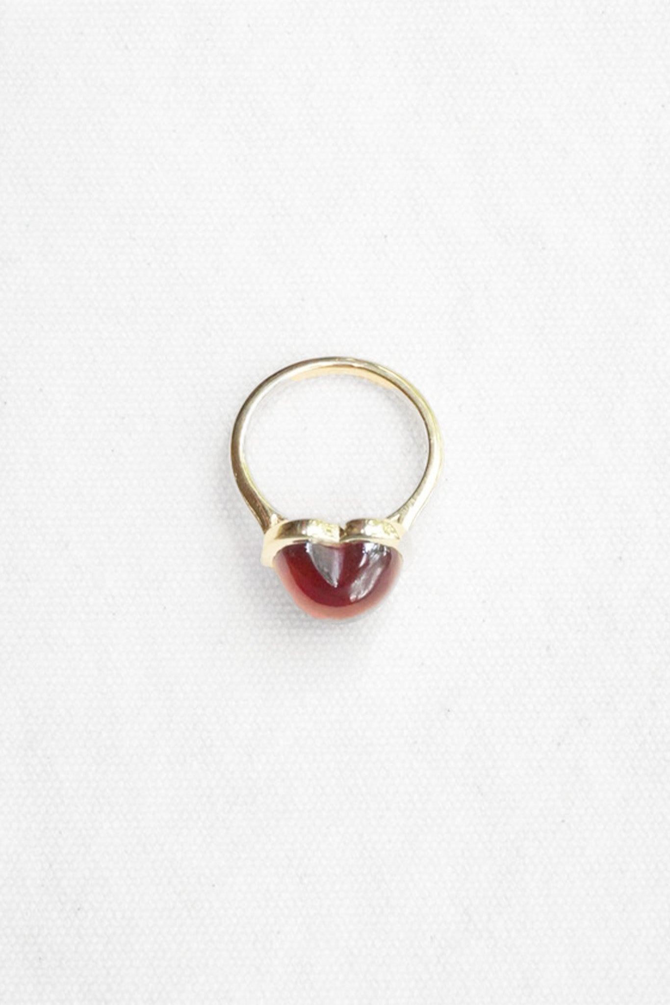 A gold ring featuring a small, polished red gemstone in a heart-shaped bezel setting, photographed on a clean white background.