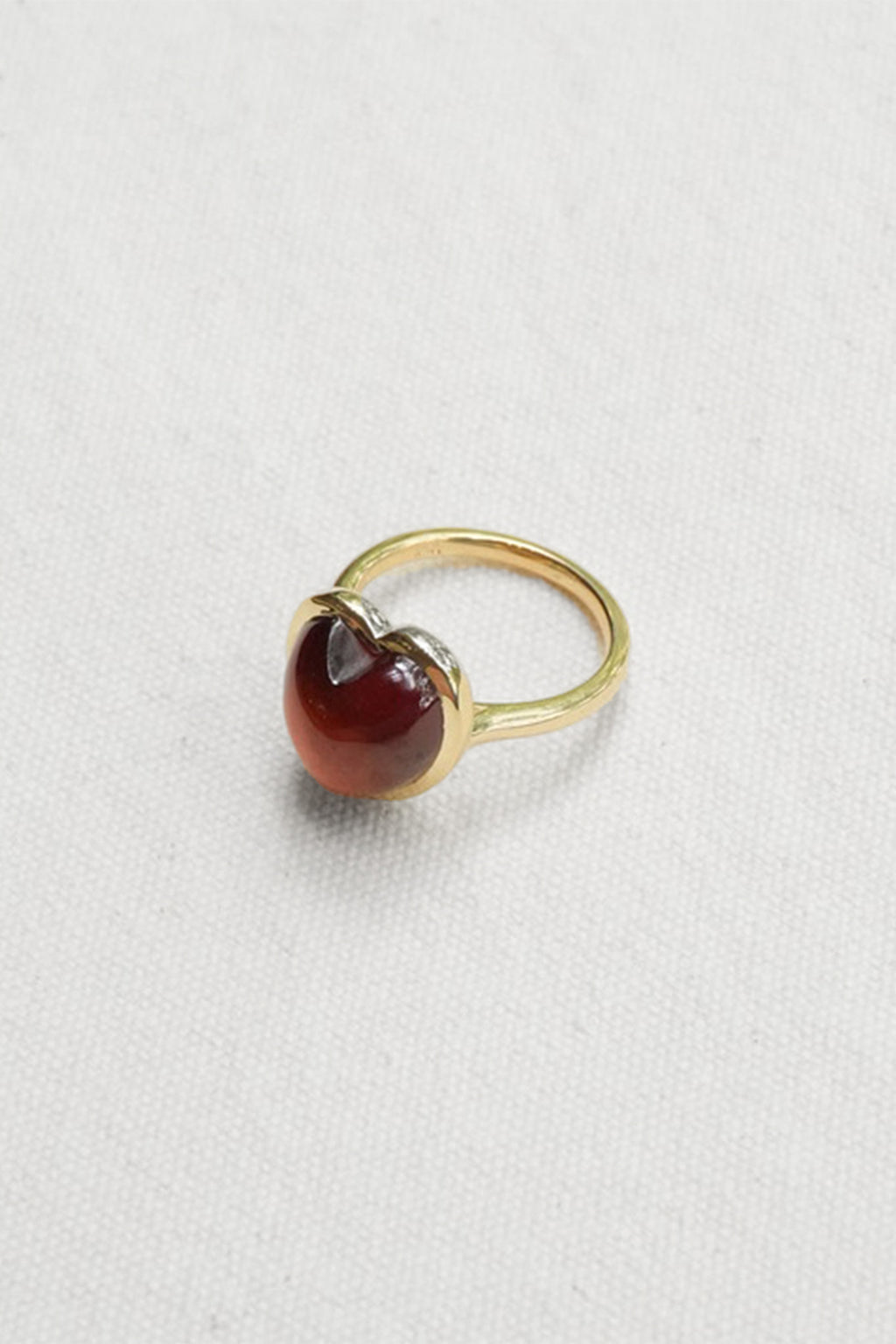 A gold ring featuring a heart-shaped cabochon gemstone in a deep reddish-brown color, set against a textured off-white fabric background.